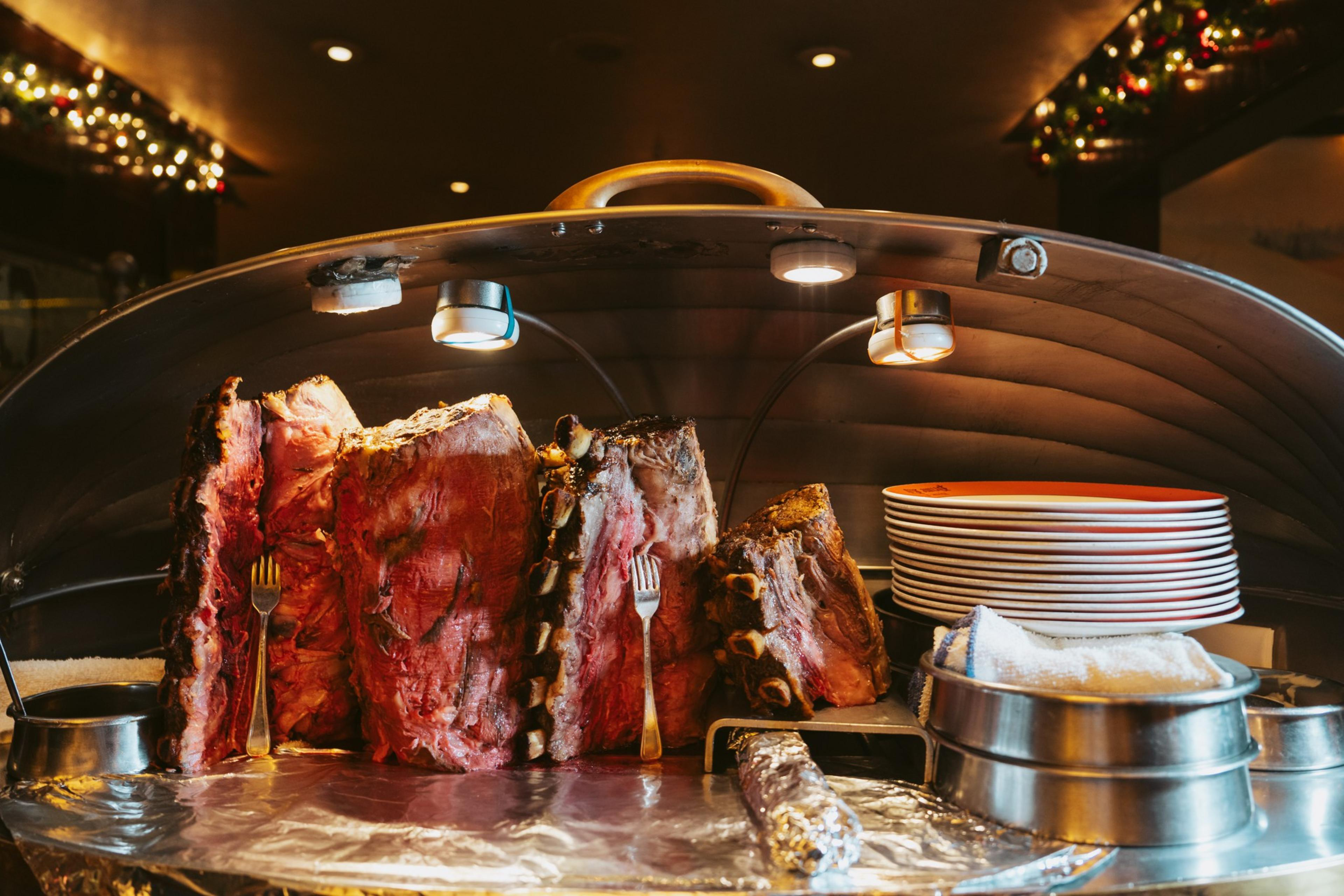 Several large, cooked beef ribs are displayed upright under a warming lamp with forks stuck in them, next to a stack of plates and a metal container.