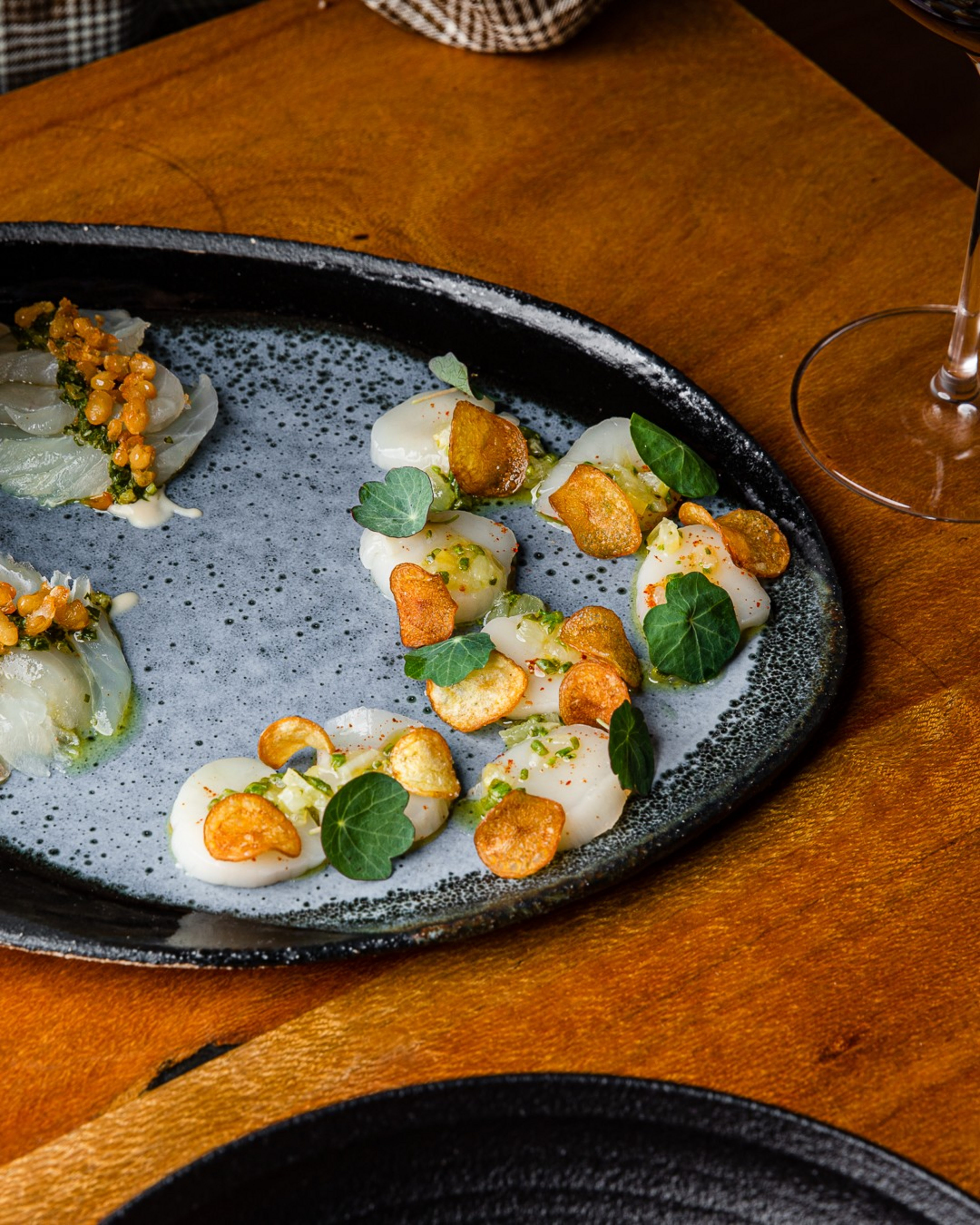 Thinly sliced scallops garnished with fried crisps, green leaves, and seasoning arranged artistically on a speckled oval plate with a glass of wine nearby.