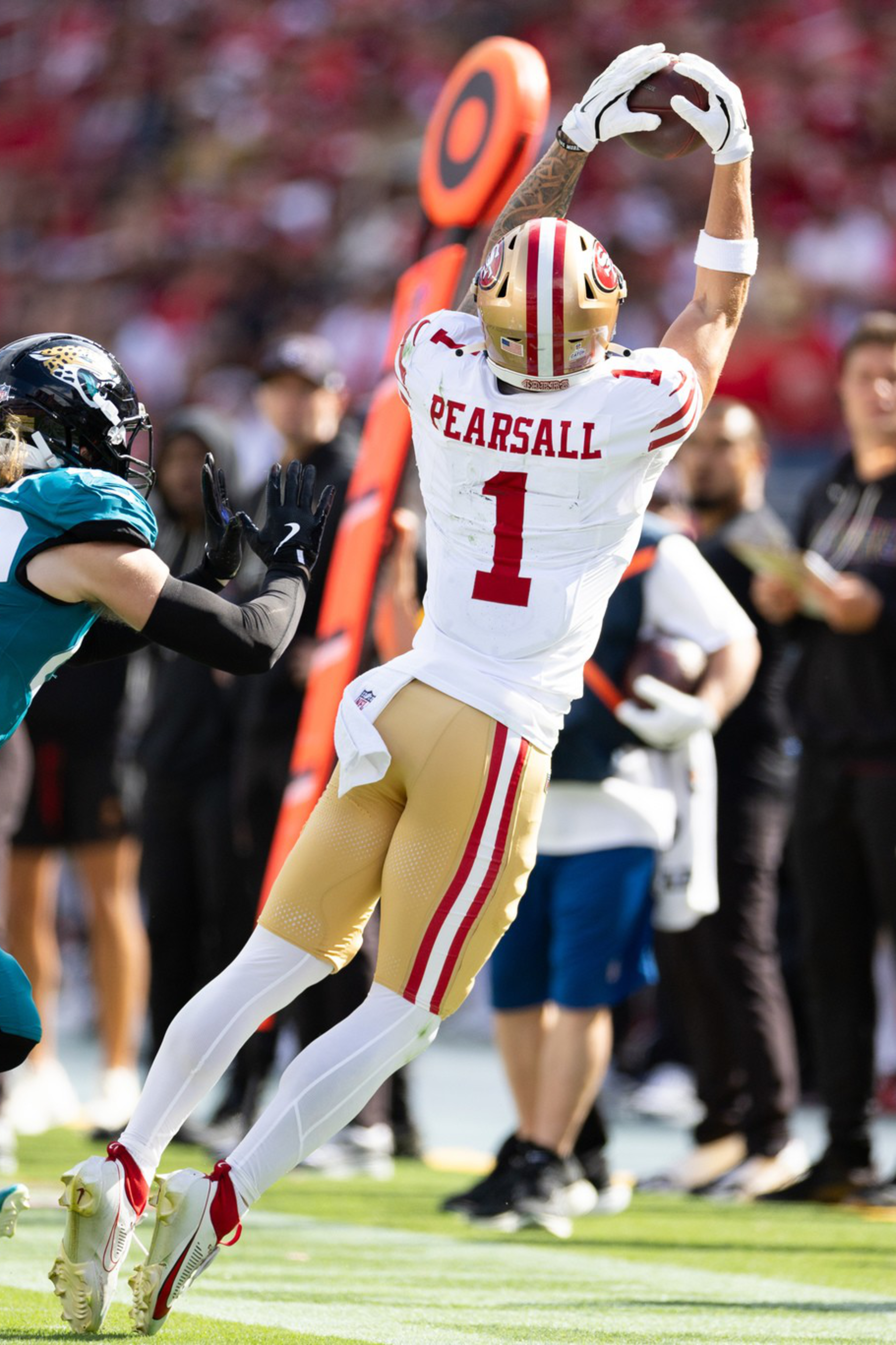 A football player in white and gold leaps to catch the ball near the sideline while two opponents in teal rush toward him.