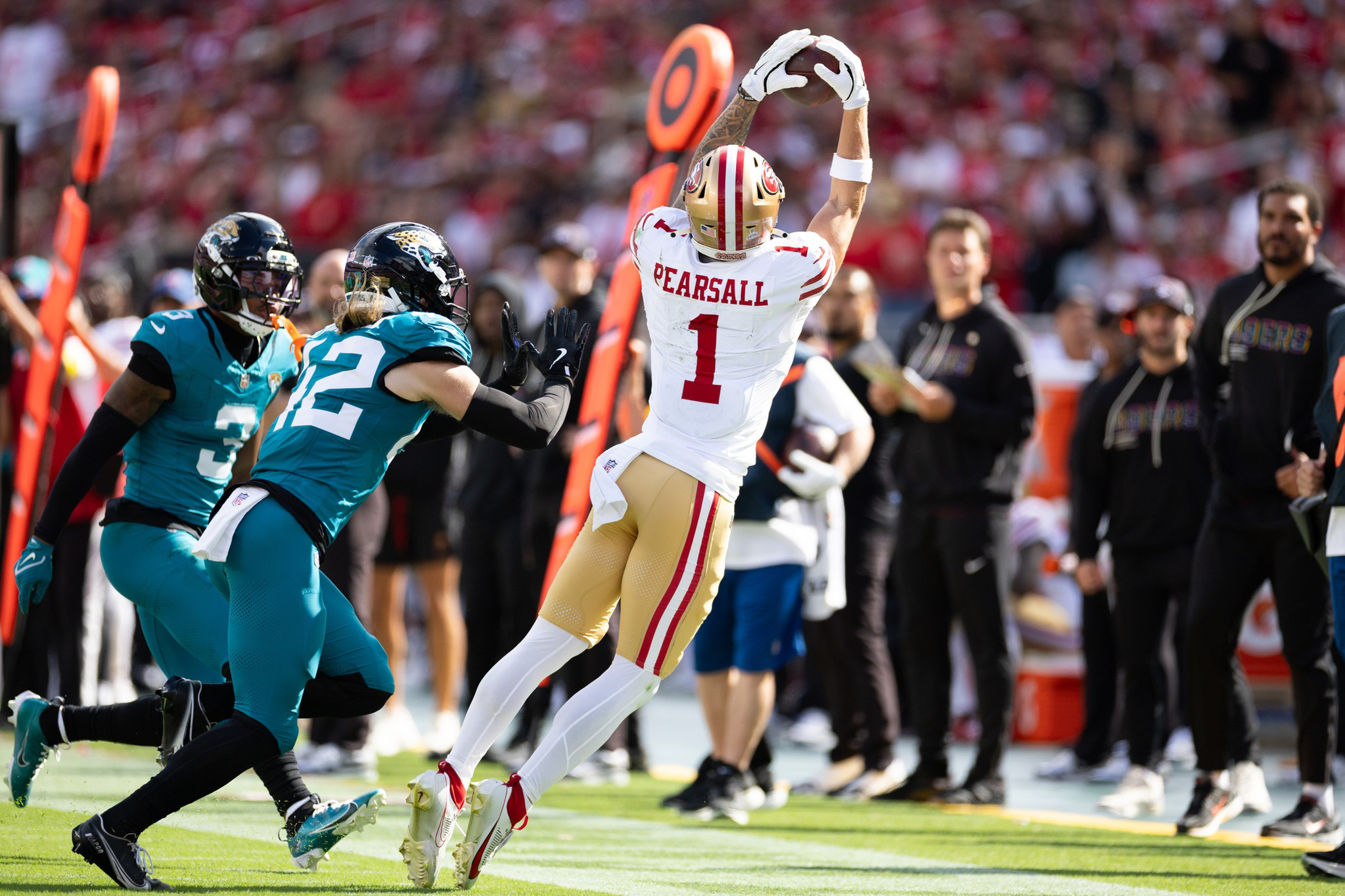 A football player in white and gold leaps to catch the ball near the sideline while two opponents in teal rush toward him.