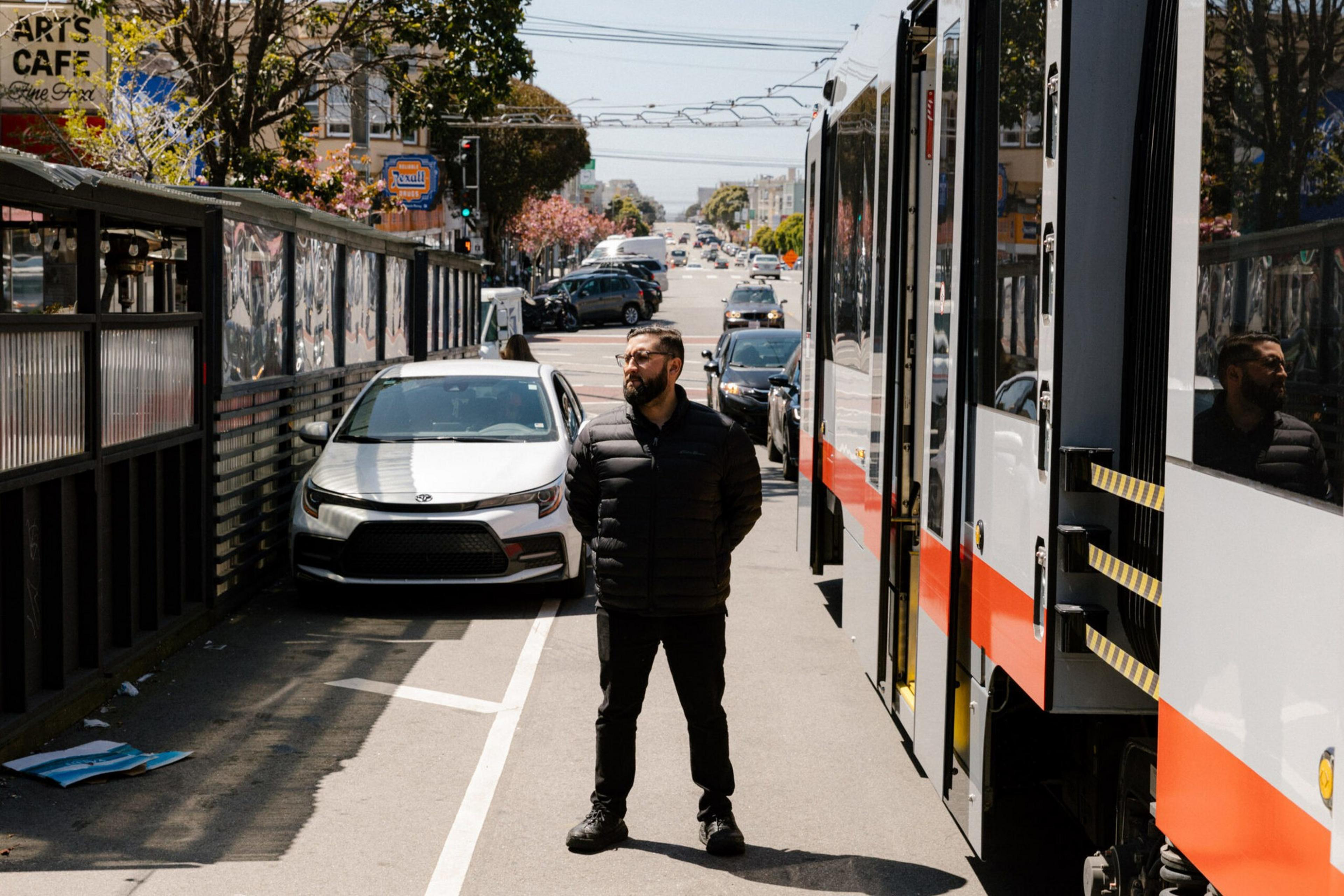 A man in black clothes and sunglasses stands on the sidewalk between a white car parked by a fence and a white and orange streetcar.
