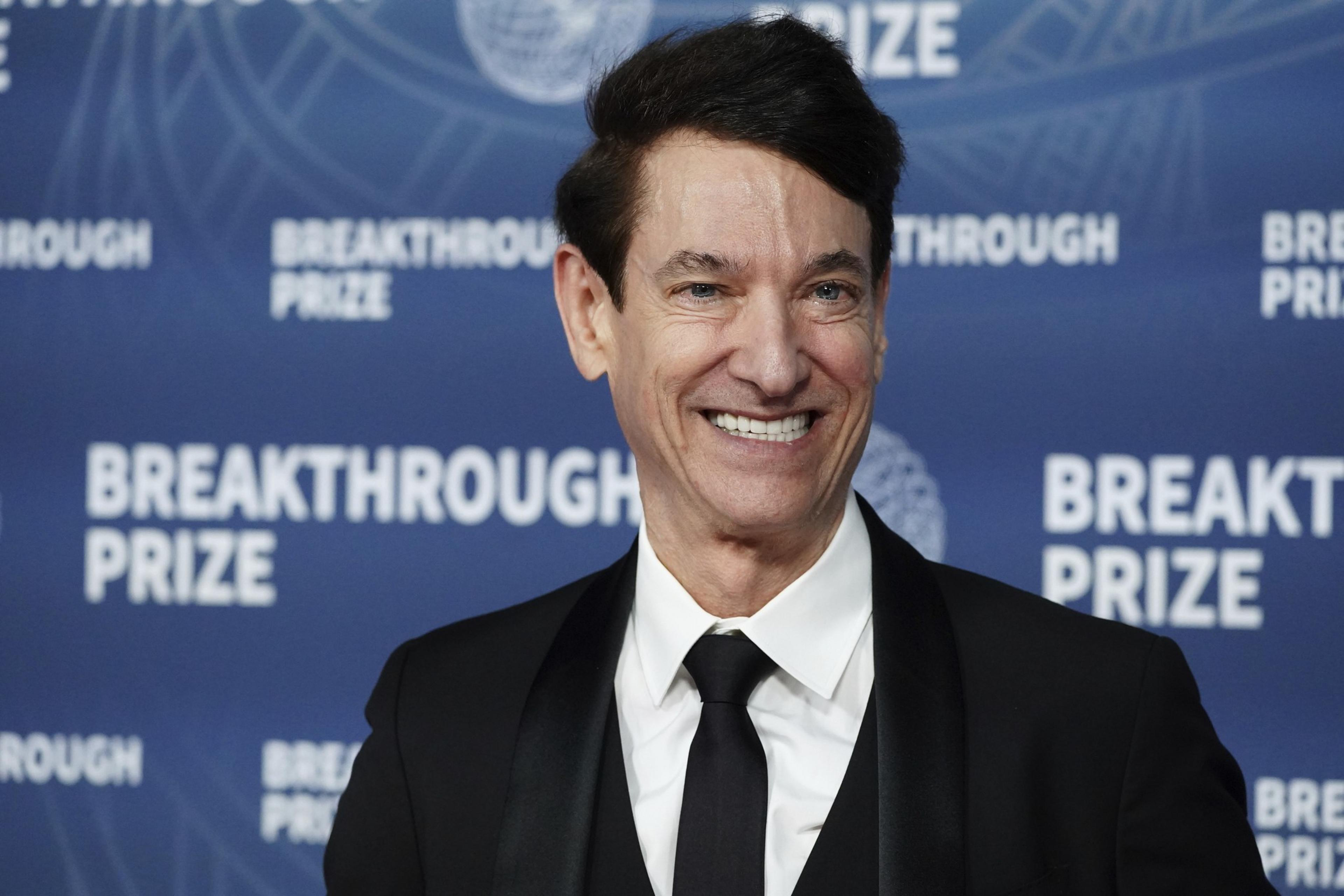 A person is smiling in front of a blue background with &quot;Breakthrough Prize&quot; repeatedly printed on it. They are wearing a black suit and tie with a white shirt.