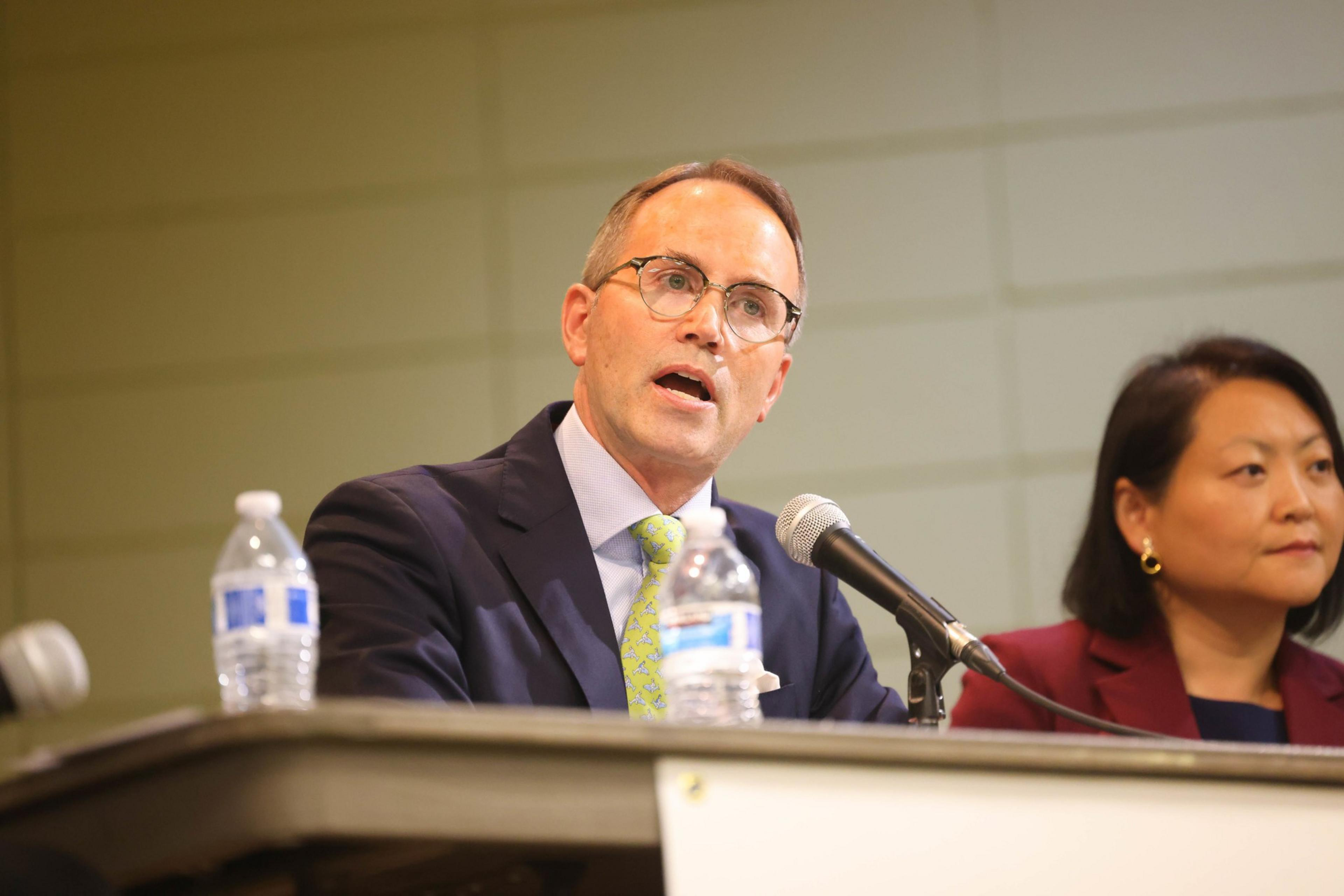 A man in glasses and a blue suit speaks into a microphone; a focused woman sits beside him, partially shown. Bottles of water are on the table.