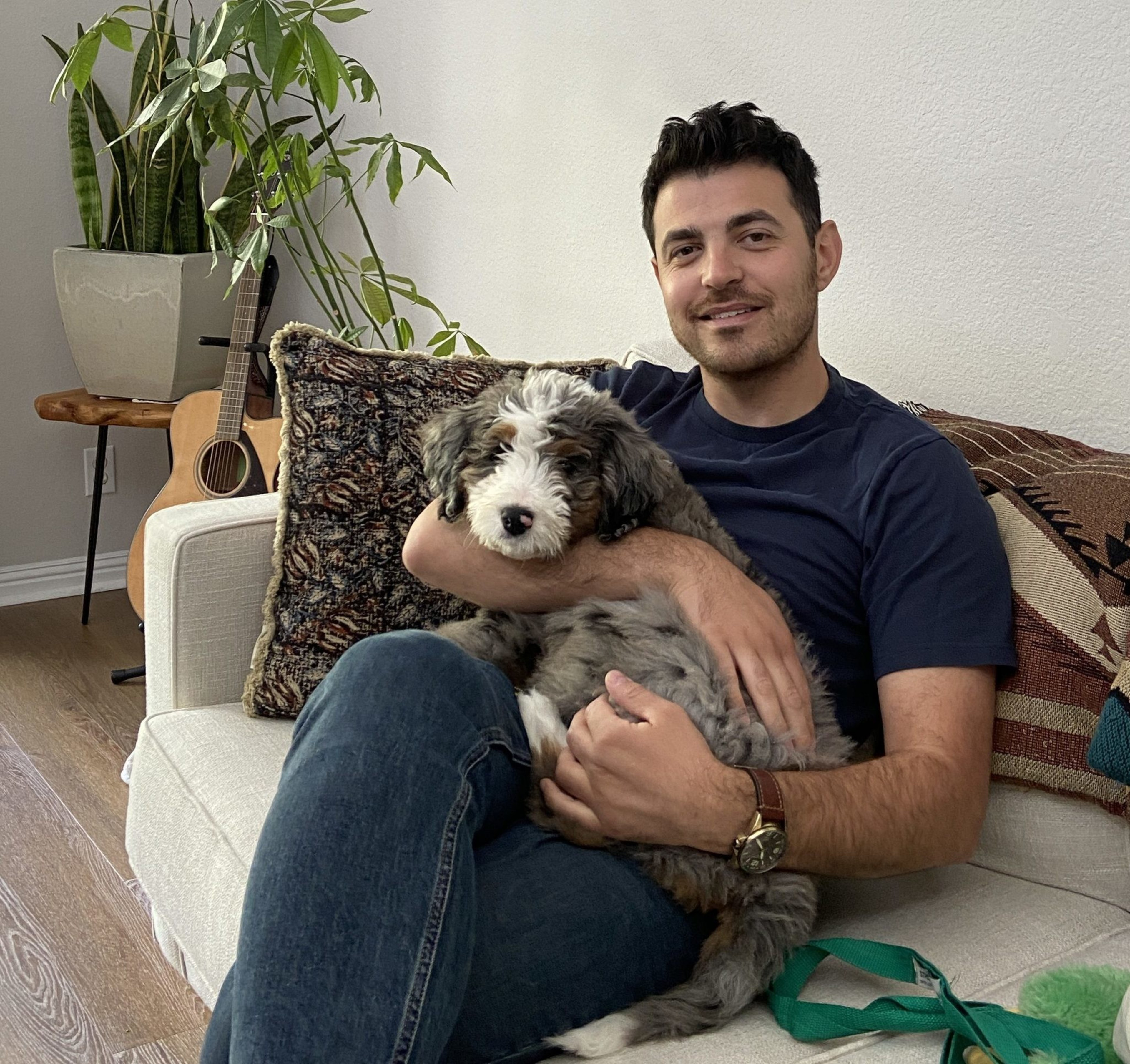A man sits on a couch holding a fluffy dog. He's wearing a dark T-shirt and jeans, with a guitar and plants in the background, creating a cozy atmosphere.