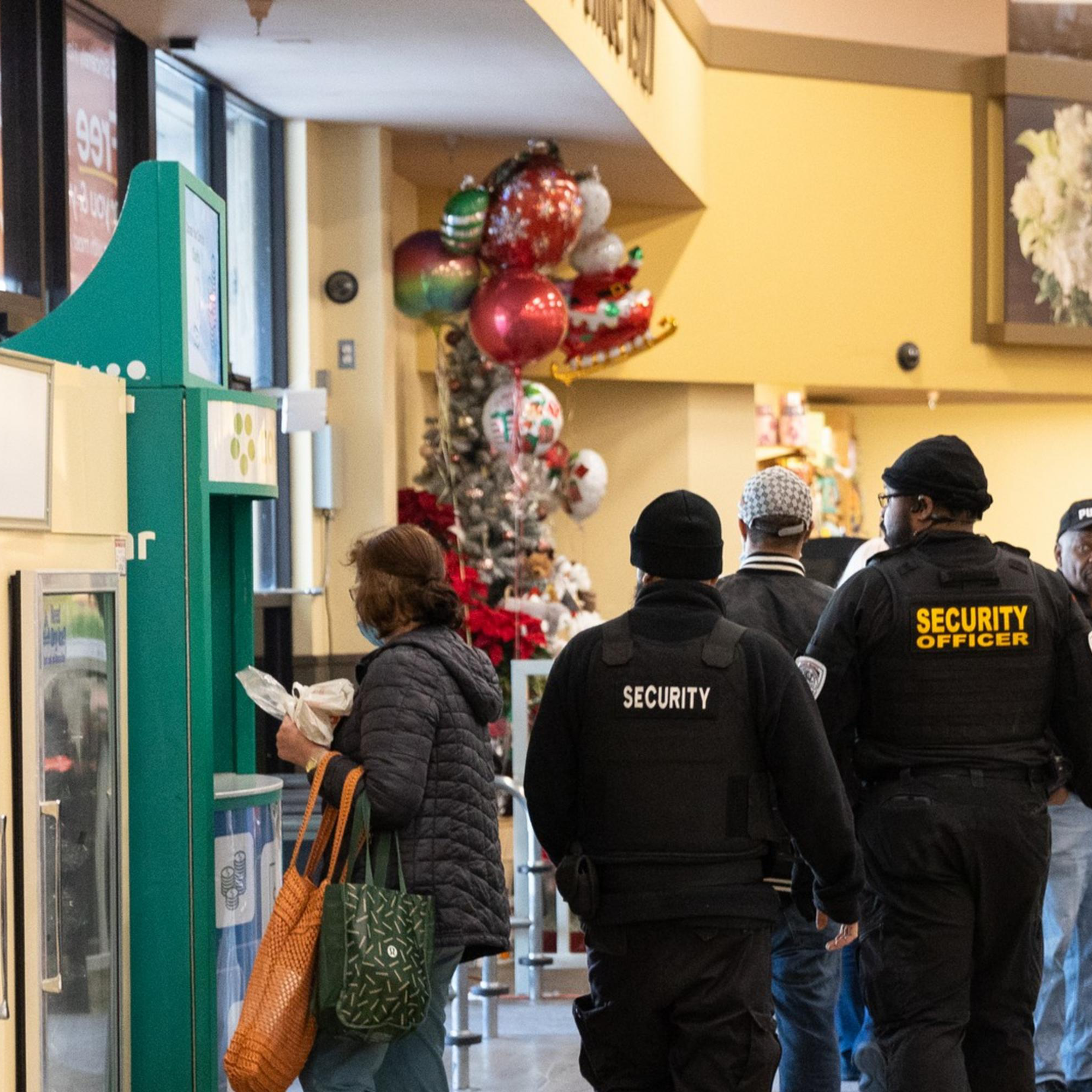 Security personnel in black uniforms walk through a brightly lit grocery store near a floral display and balloons, while a customer stands nearby.