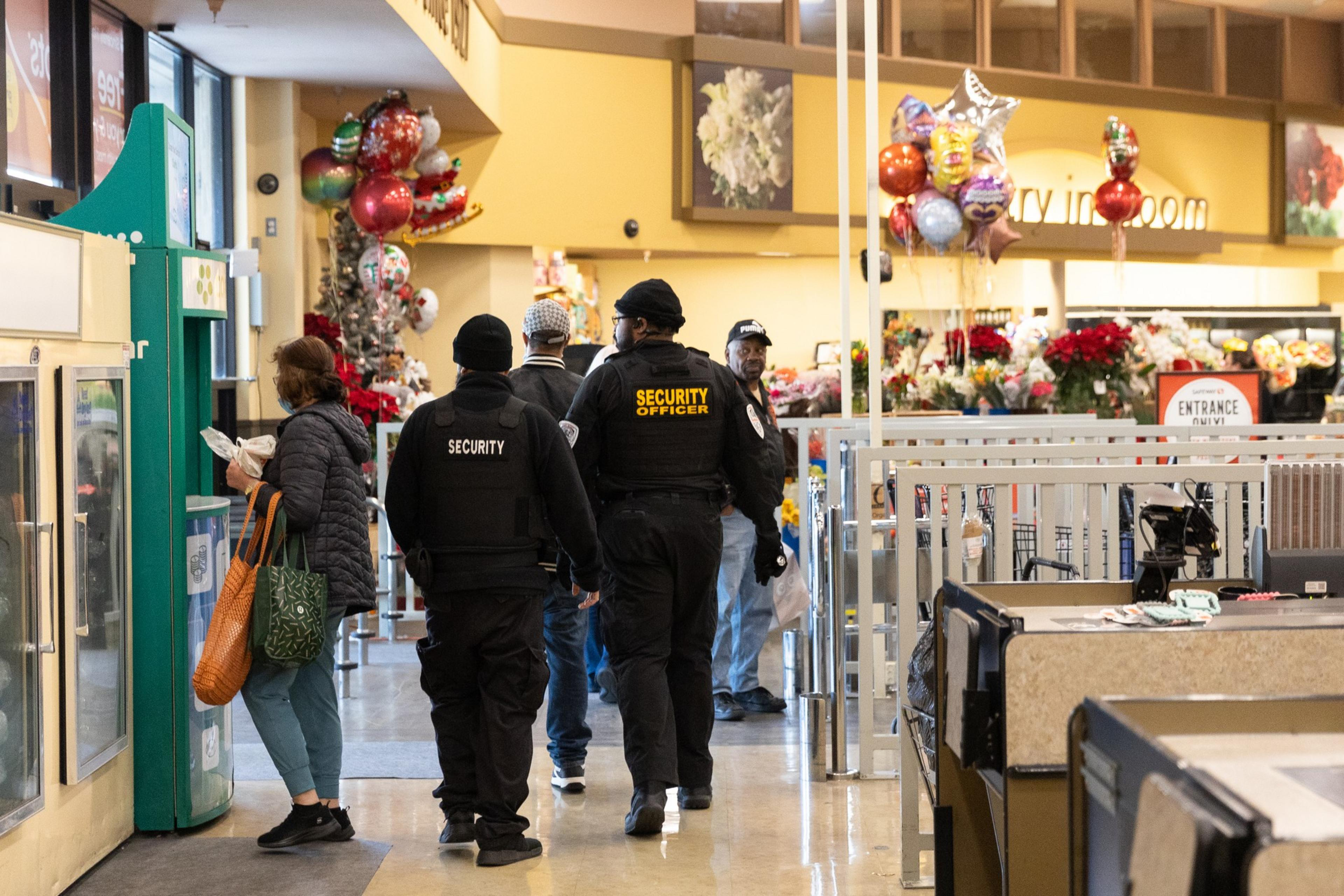 Security personnel in black uniforms walk through a brightly lit grocery store near a floral display and balloons, while a customer stands nearby.