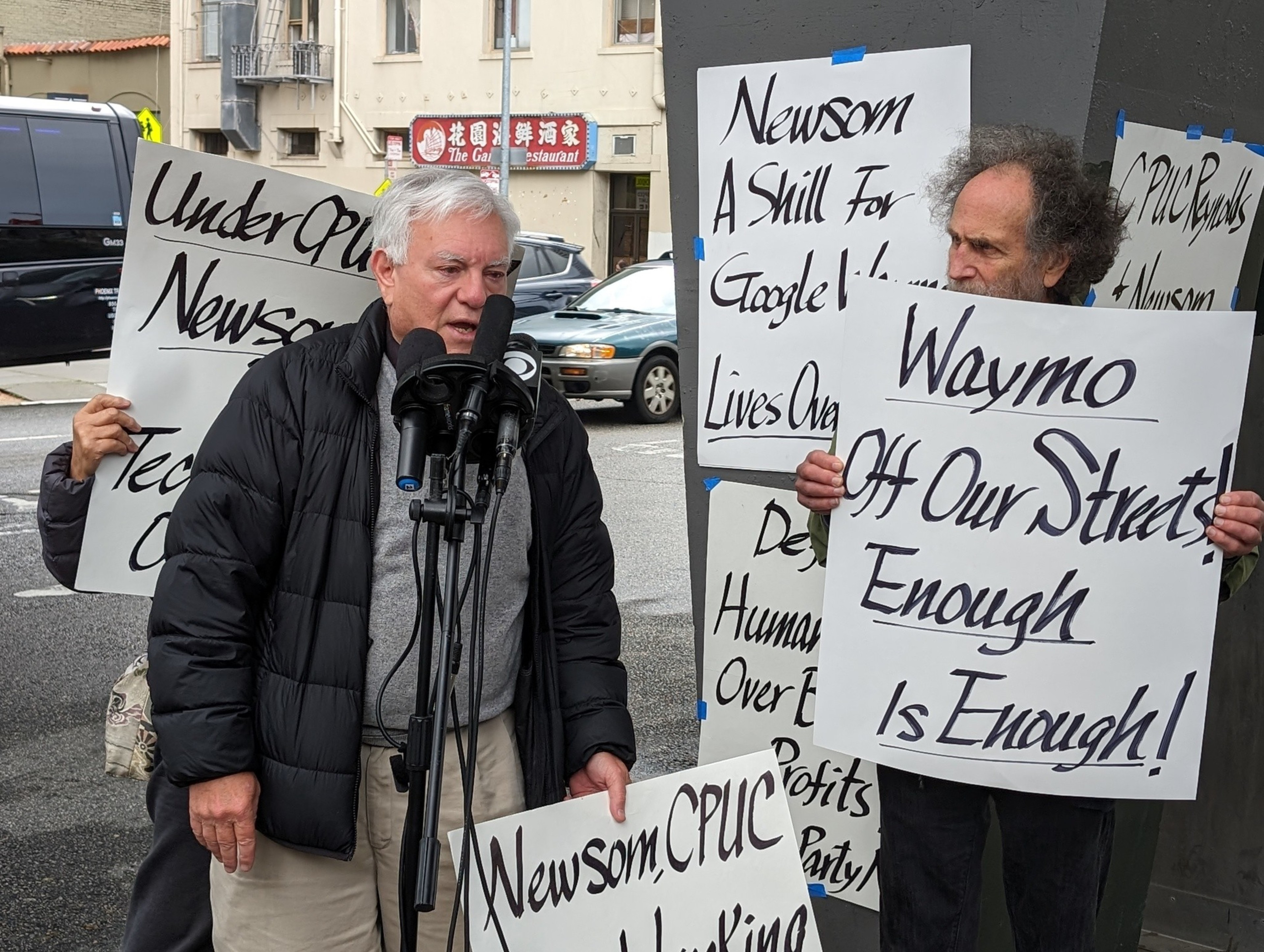 A man holds a posterboard sign and stands next to another speaking into a stand for multiple microphones.