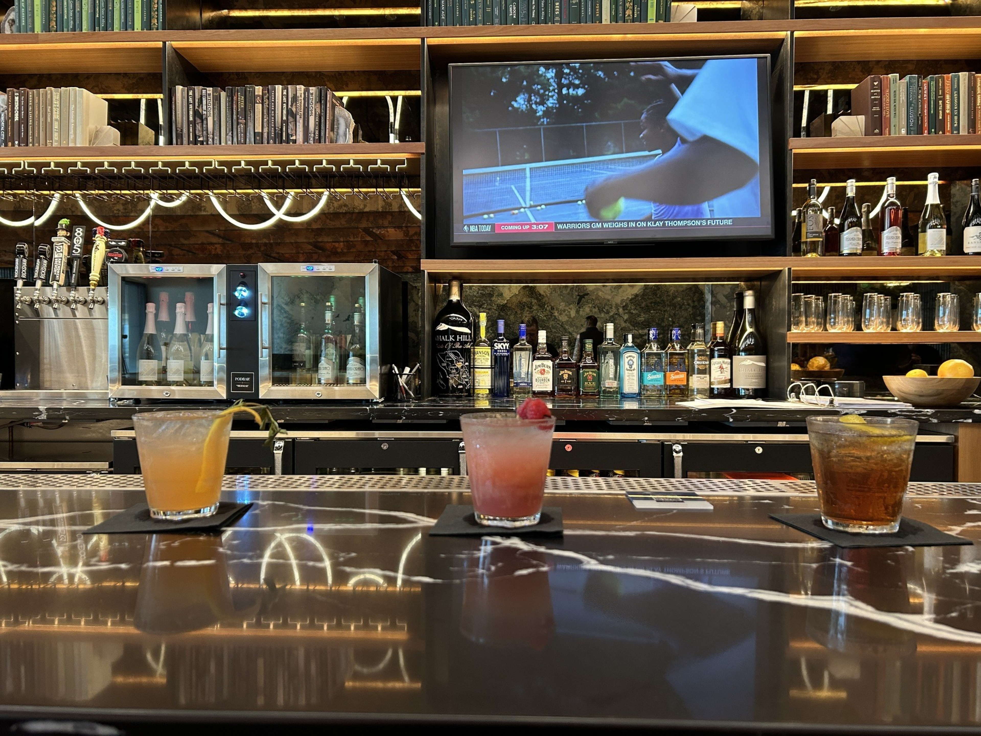 The image shows a bar counter with three cocktails, backed by a display of liquor bottles, a wine cooler, beer taps, and a wall-mounted TV playing a tennis match.
