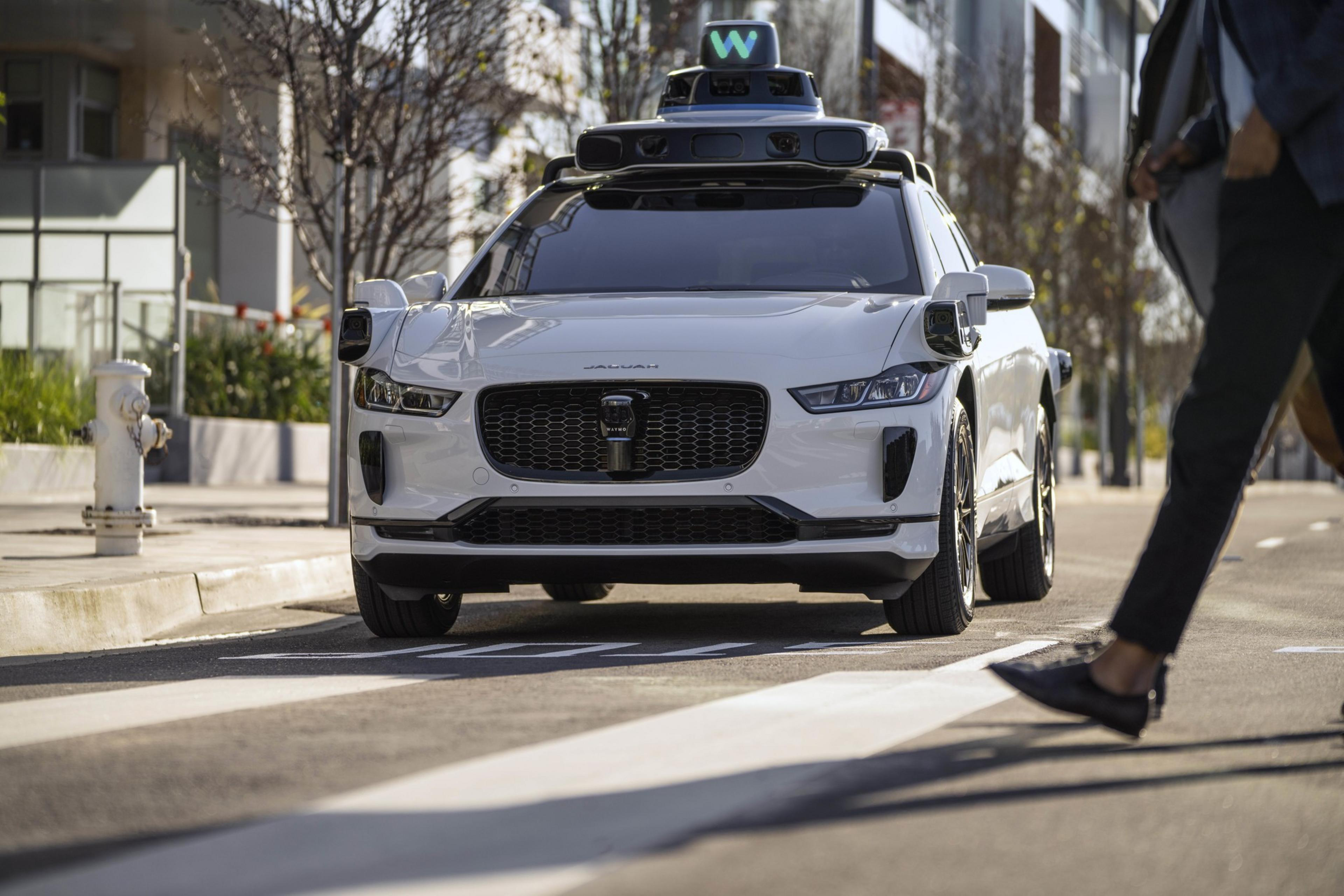 A white self-driving car with sensors on top stops at a crosswalk as a pedestrian walks by. It's on a city street lined with modern buildings and trees.