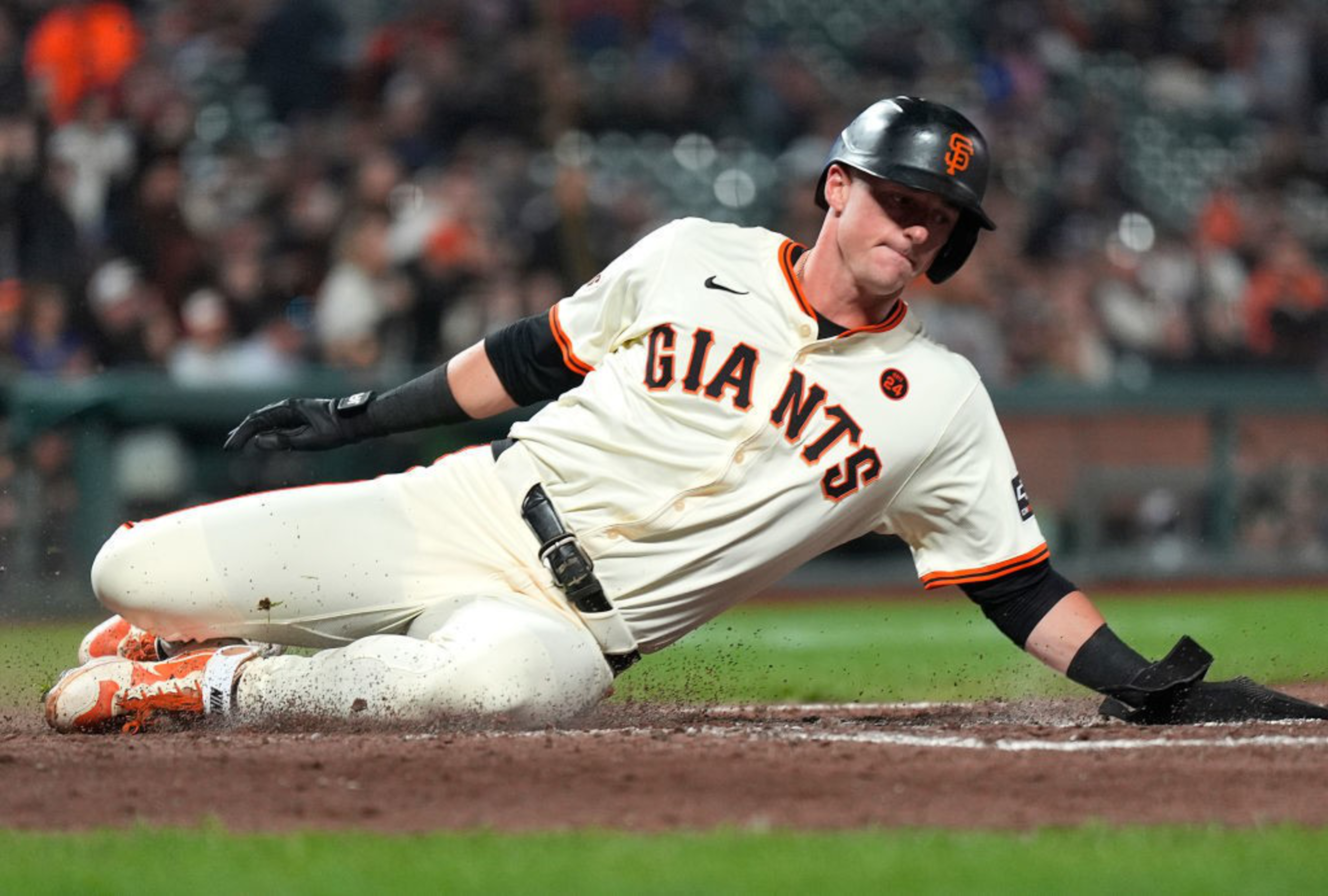 A baseball player wearing a white "Giants" uniform and helmet slides into a base on a dirt field, with a focused expression during a game.