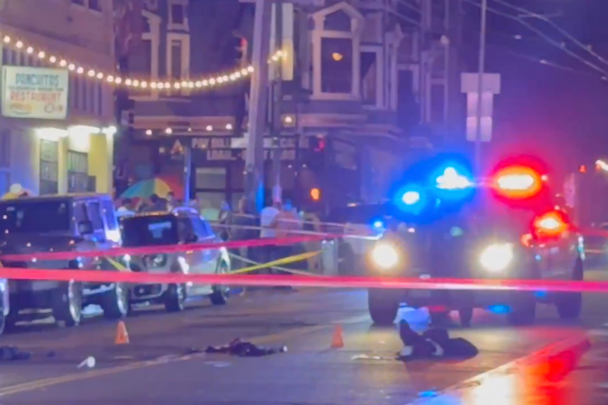 A nighttime street scene with police vehicles flashing red and blue lights, red police tape blocking the area, and several small evidence markers on the ground.