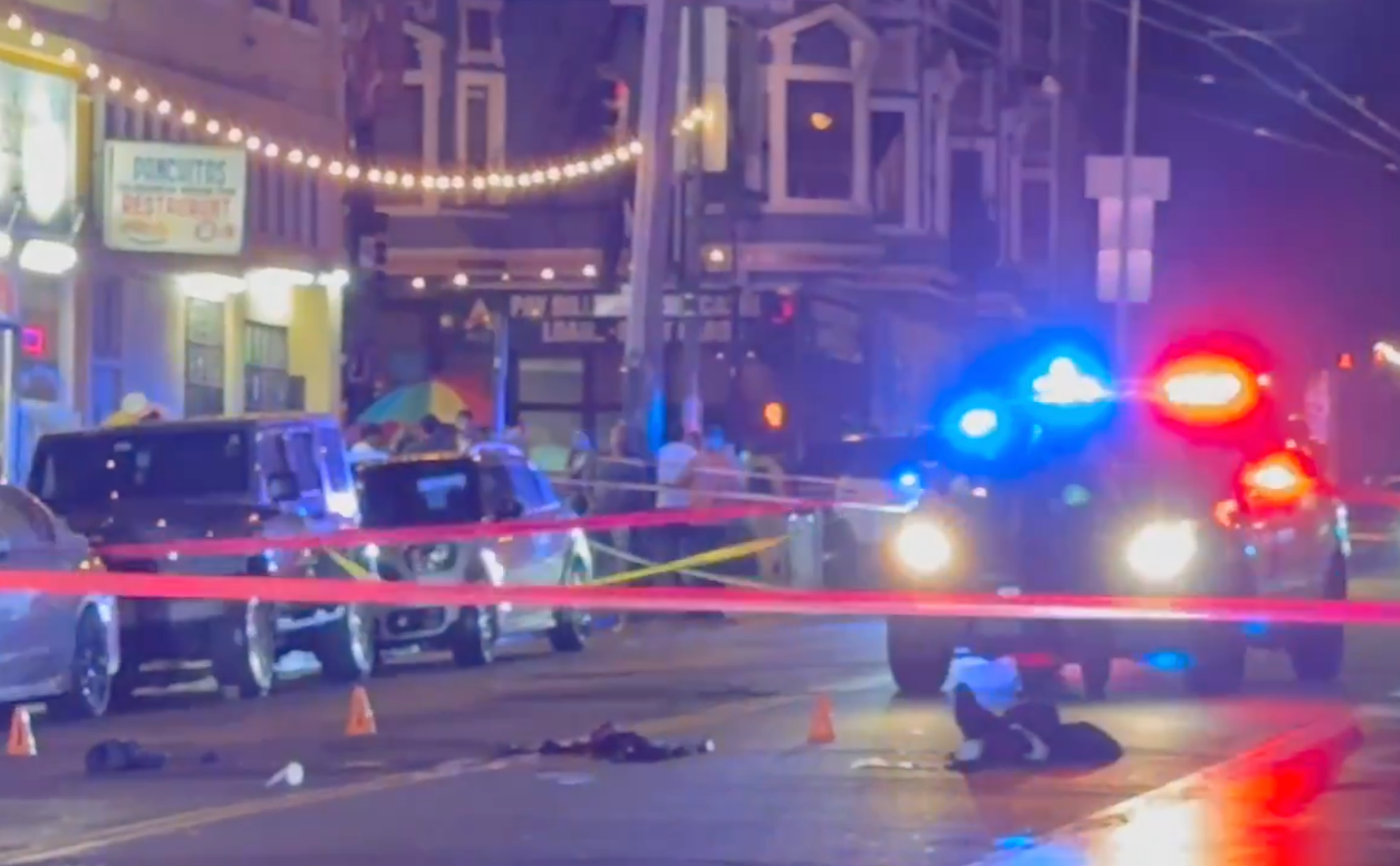 A nighttime street scene with police vehicles flashing red and blue lights, red police tape blocking the area, and several small evidence markers on the ground.