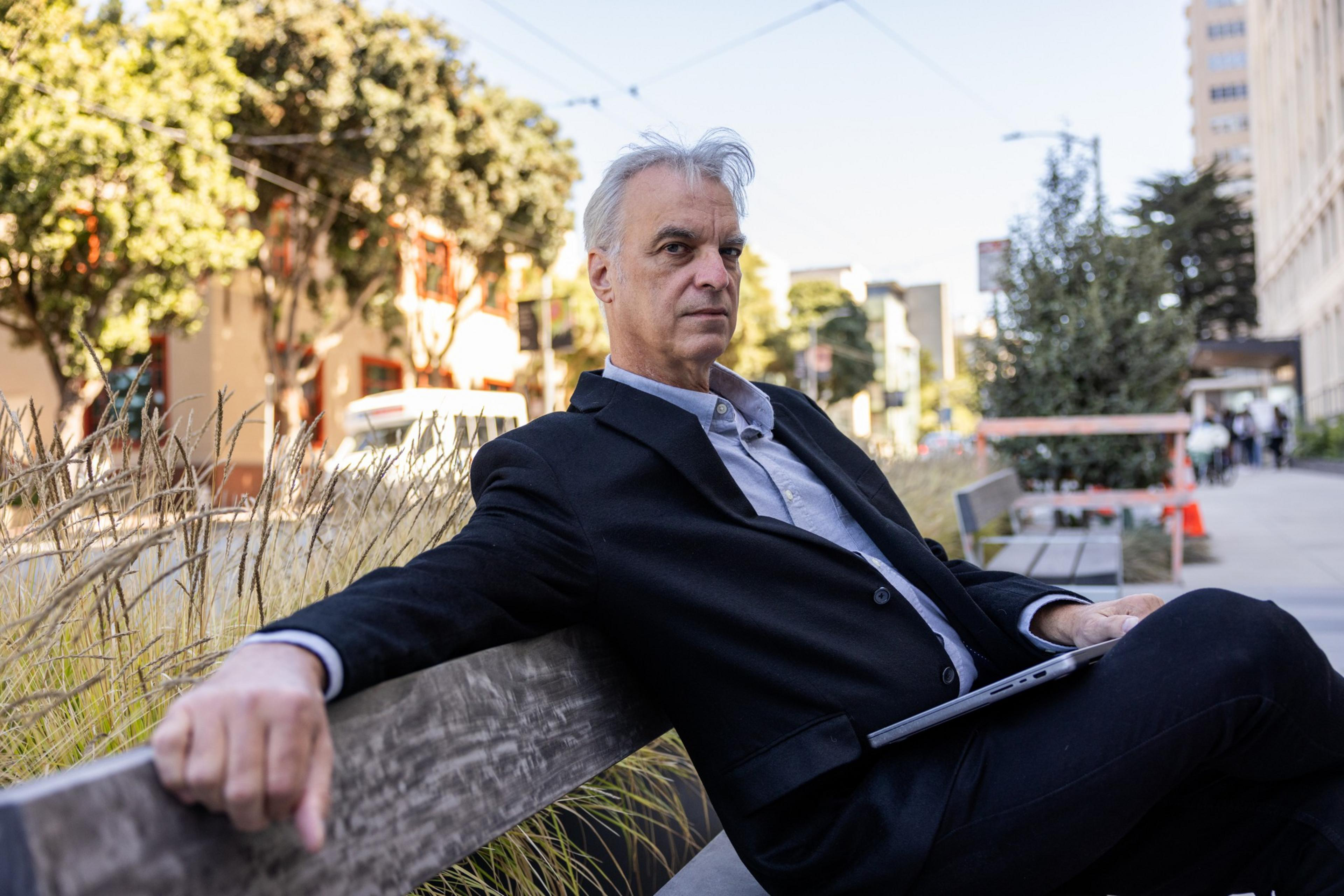 A man with gray hair in a black suit sits on a wooden bench outdoors, holding a closed laptop with trees and buildings in the background.
