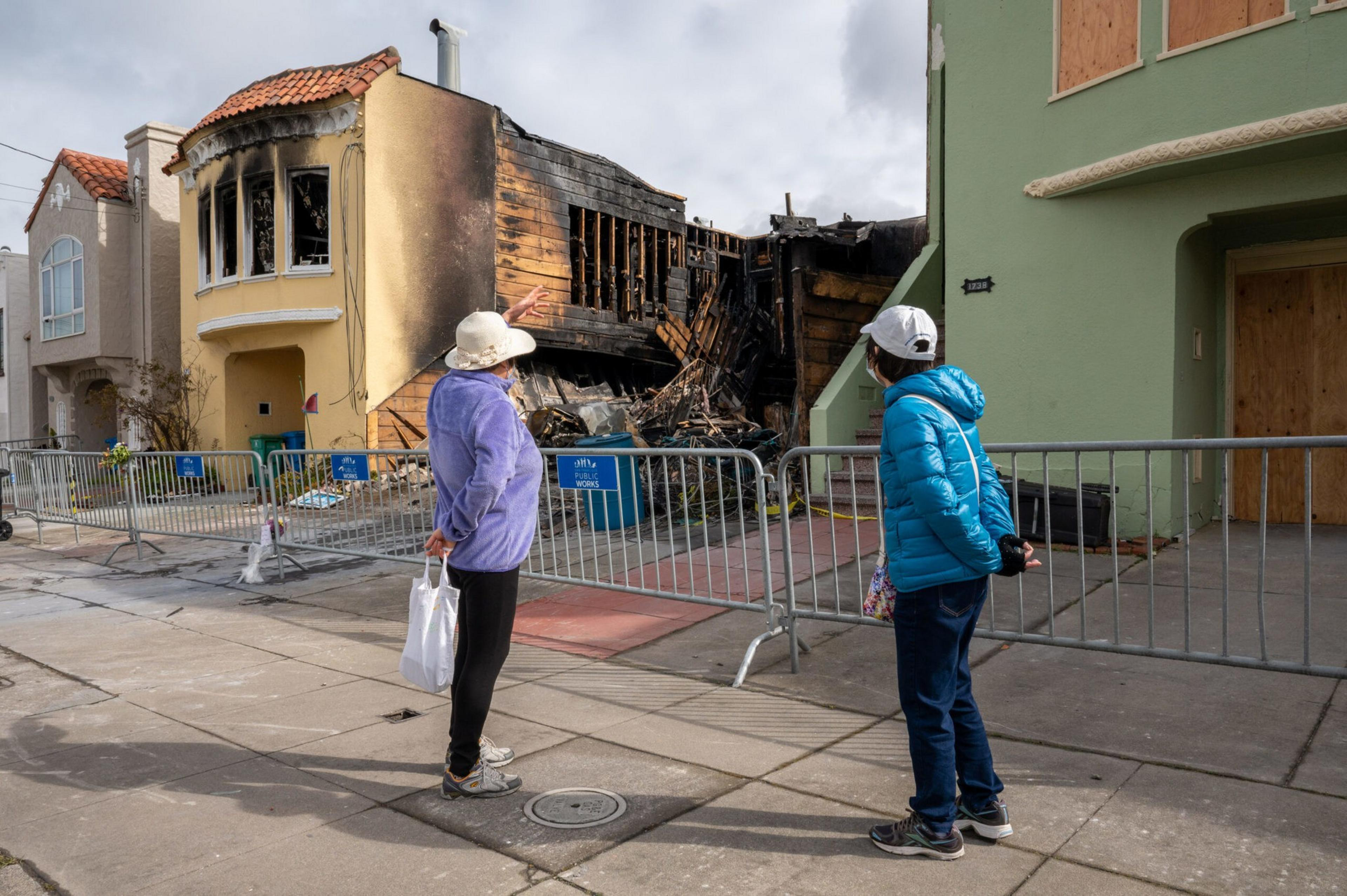 Two people stand behind metal barricades looking at a severely fire-damaged house with charred walls and debris.