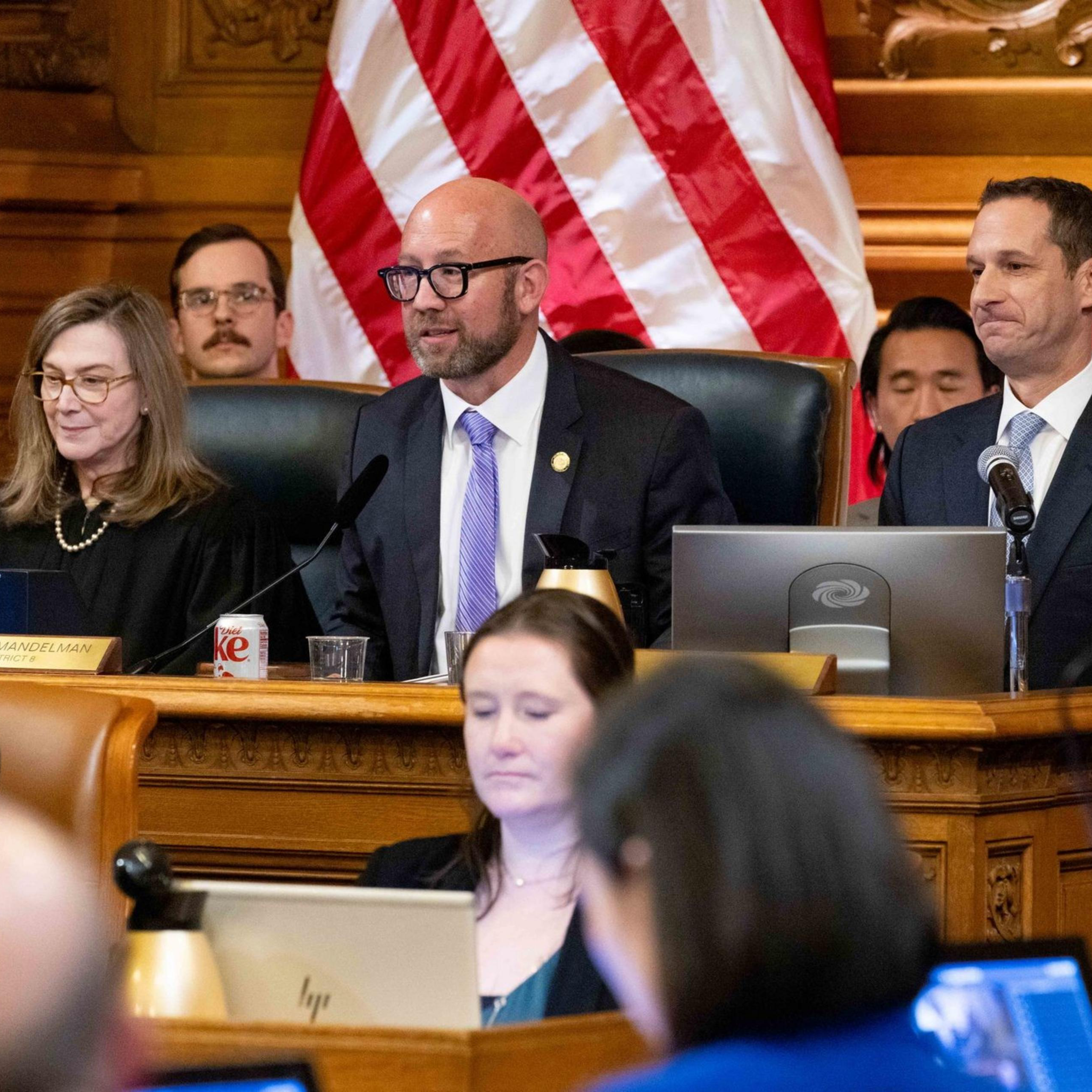Several people are seated in a formal setting with microphones and flags in the background, suggesting a government or official meeting. They appear to be engaged in discussion.
