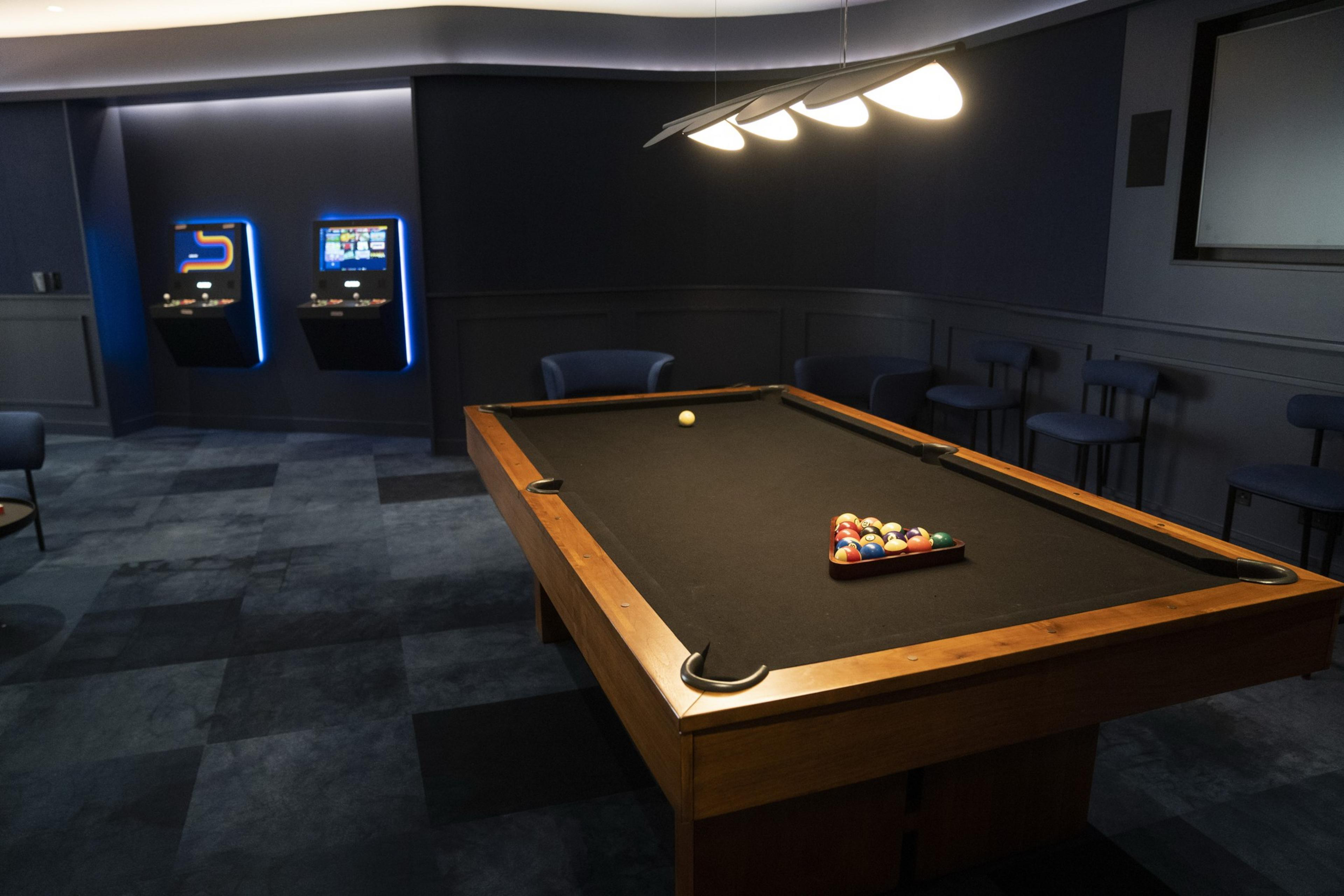 A modern game room features a pool table with balls arranged. Two arcade machines glow in the corner, and several bar stools are placed against dark-paneled walls.