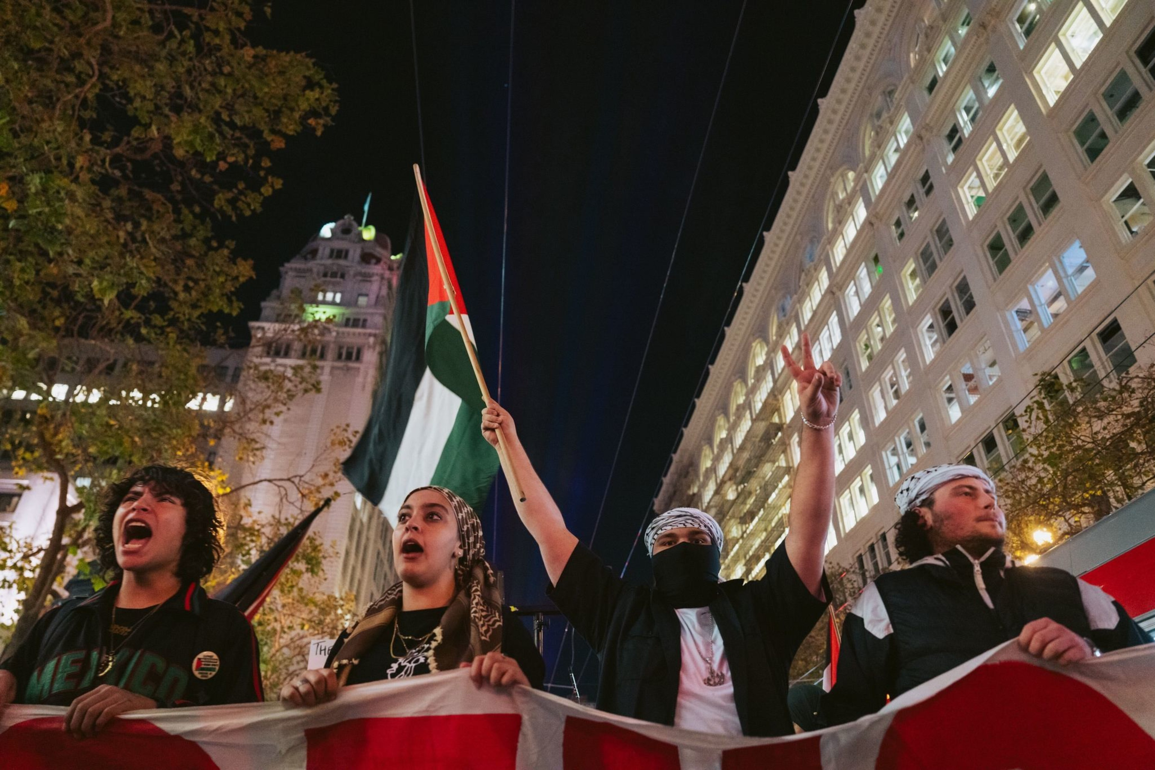 Four protesters holding a banner, with one of them wearing a mask and raising a Palestinian flag. 