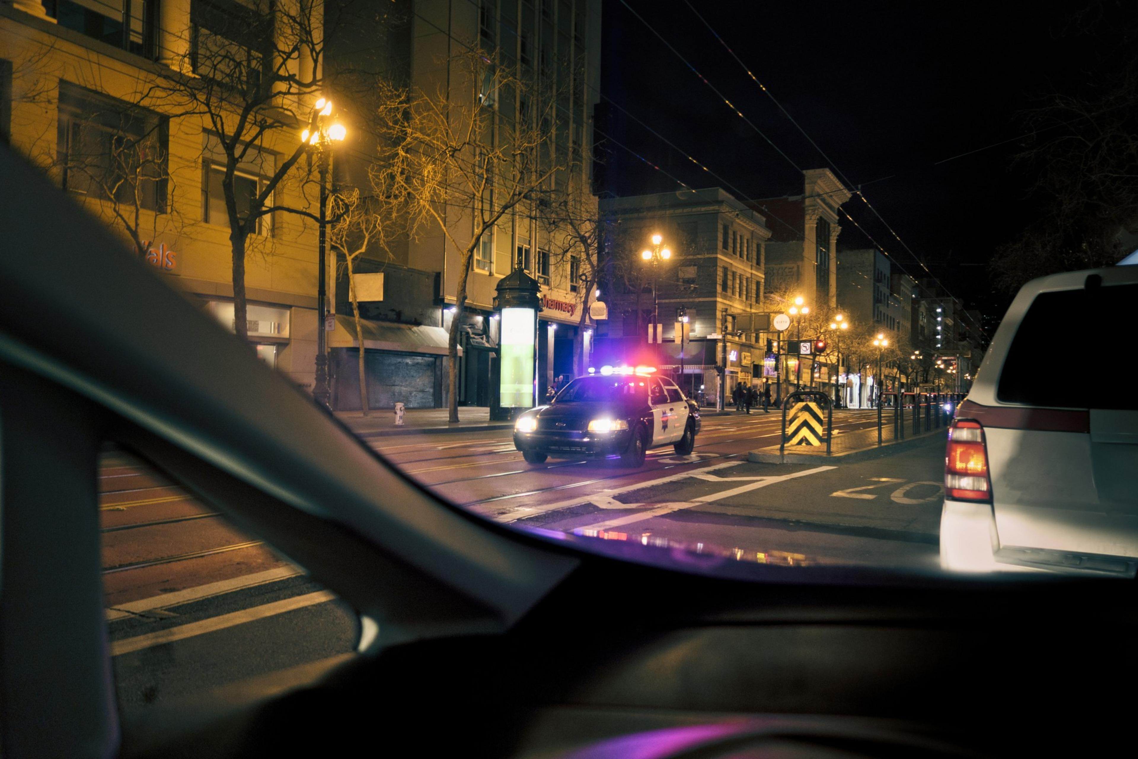 In the photo, there's a nighttime urban street scene viewed from a car. A police car with flashing lights is in the center, and buildings with lit windows line the street.