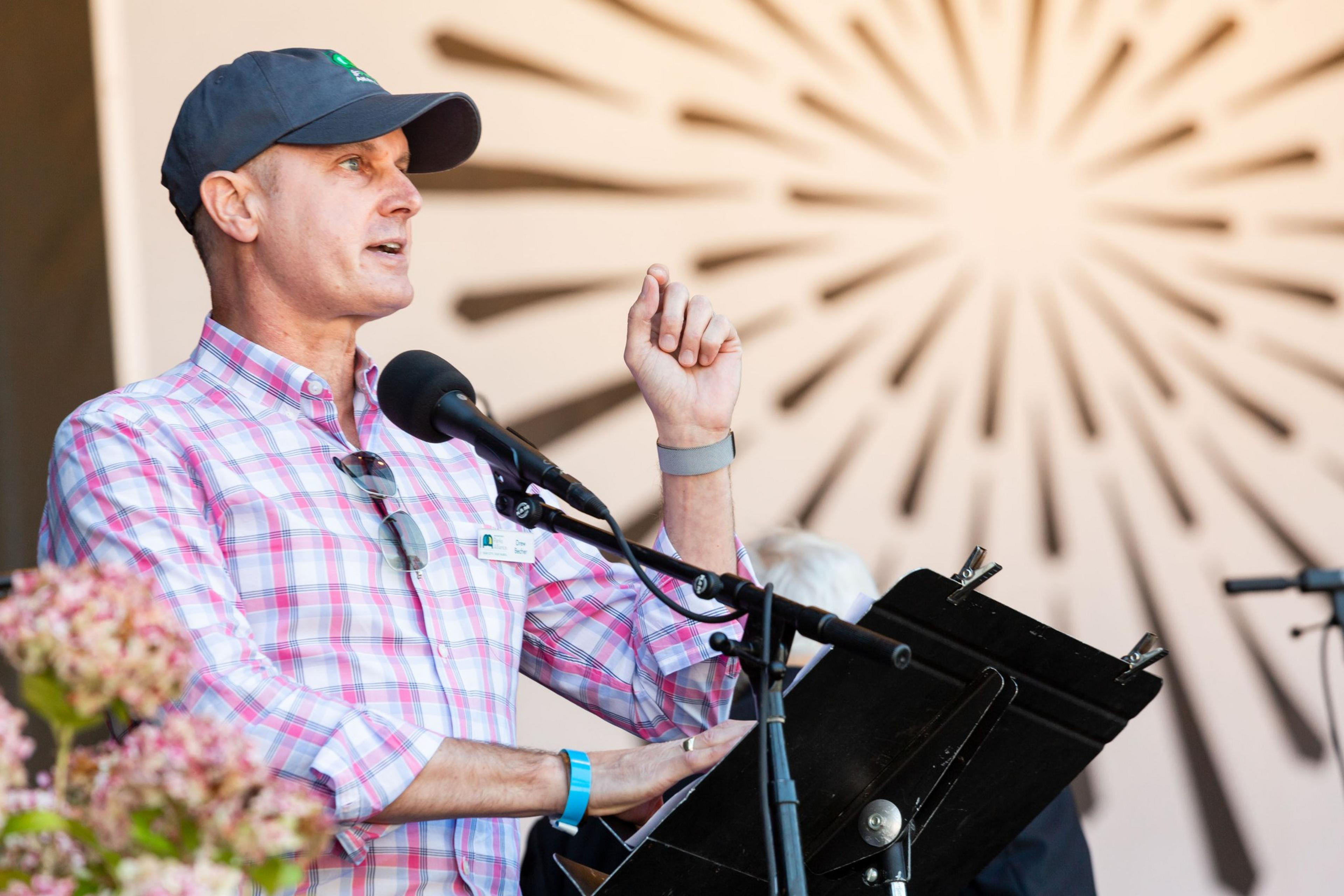 A person in a plaid shirt and cap speaks at a microphone on a stage, gesturing with one hand. Bright rays are depicted on a background wall behind him.