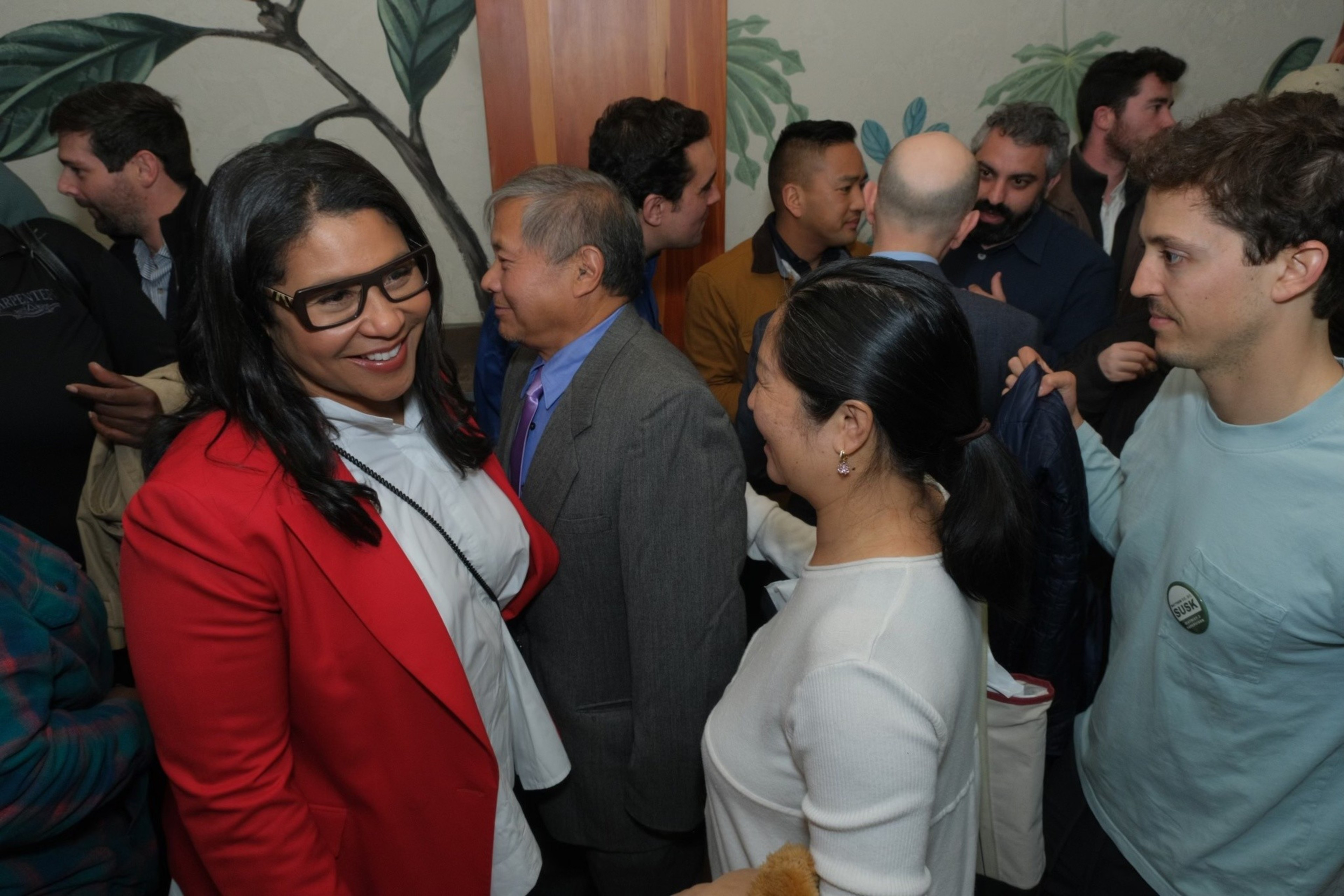 A crowded room with diverse people conversing, two women in the foreground smiling at each other.