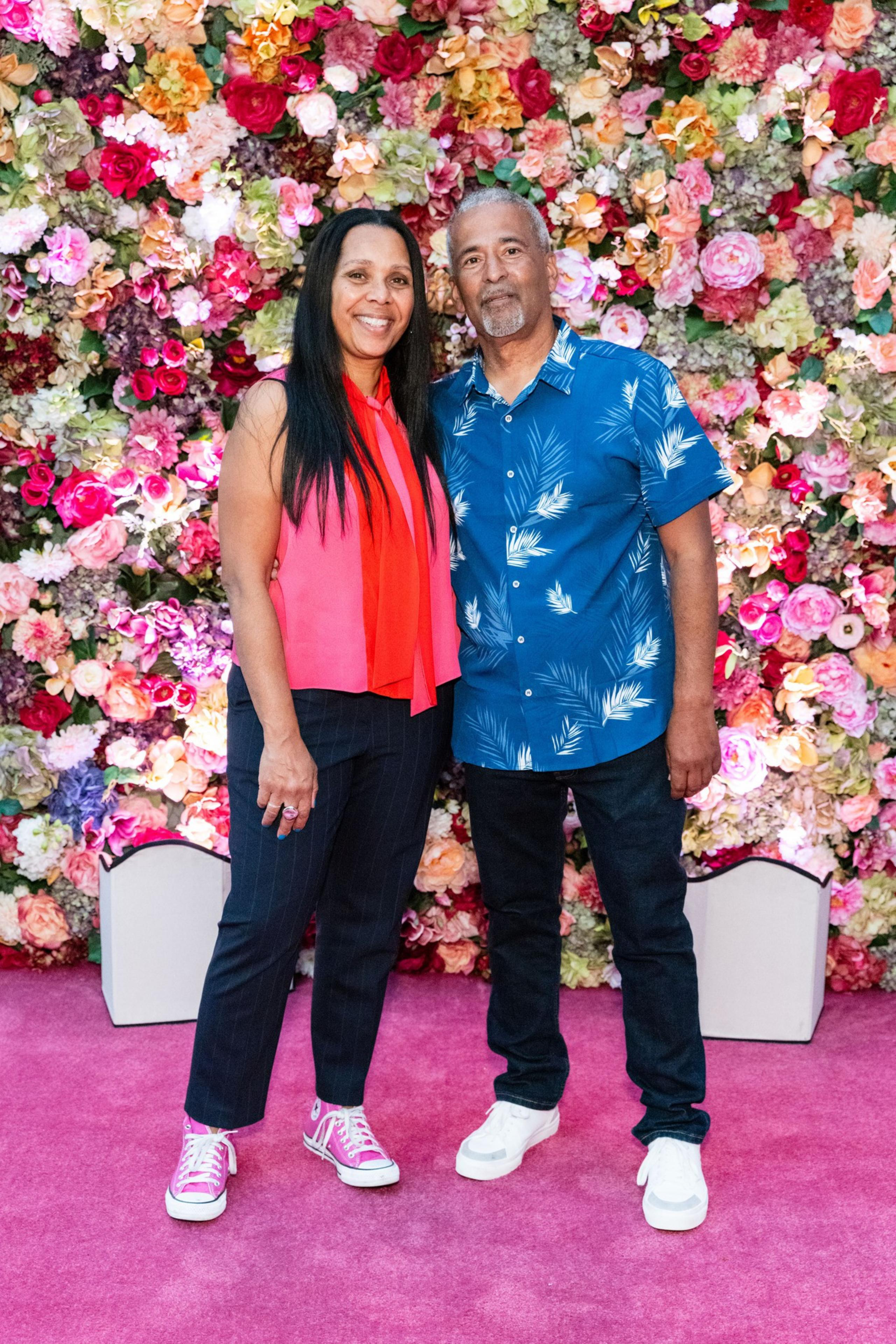 A smiling couple stands on a pink carpet, with a vibrant floral wall behind them. The woman wears a red top and pink sneakers, and the man sports a blue tropical shirt.