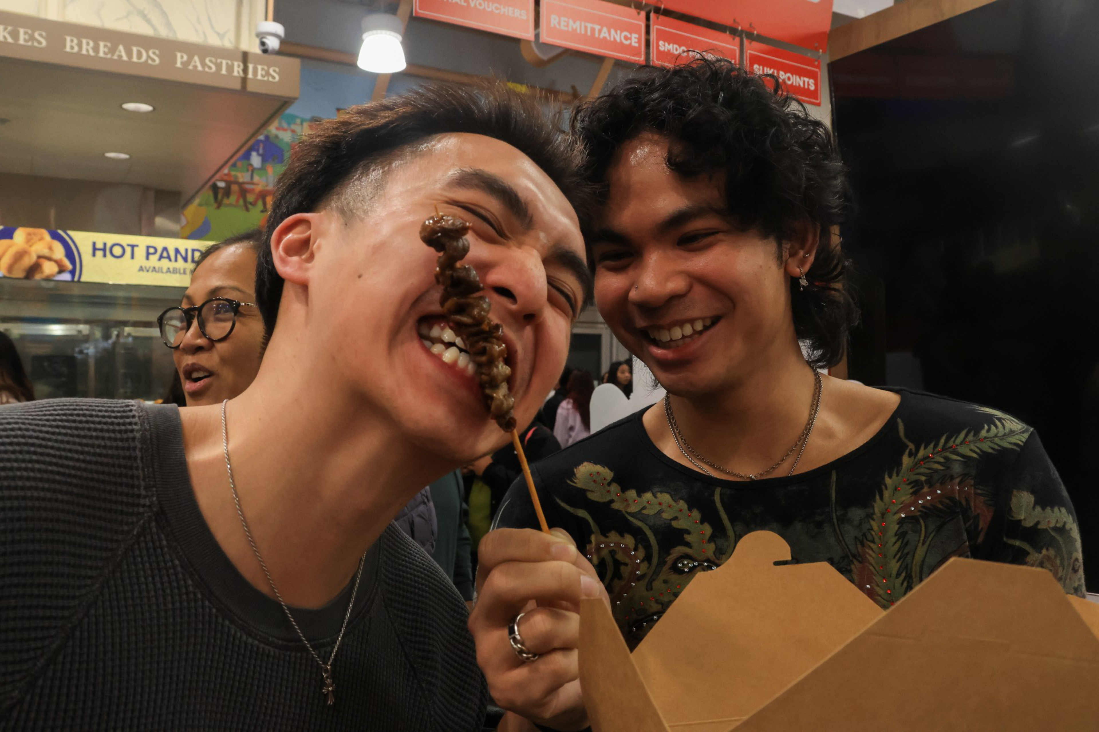 A man bites a grilled meat skewer while another smiles and holds a takeout box inside a busy indoor food market.