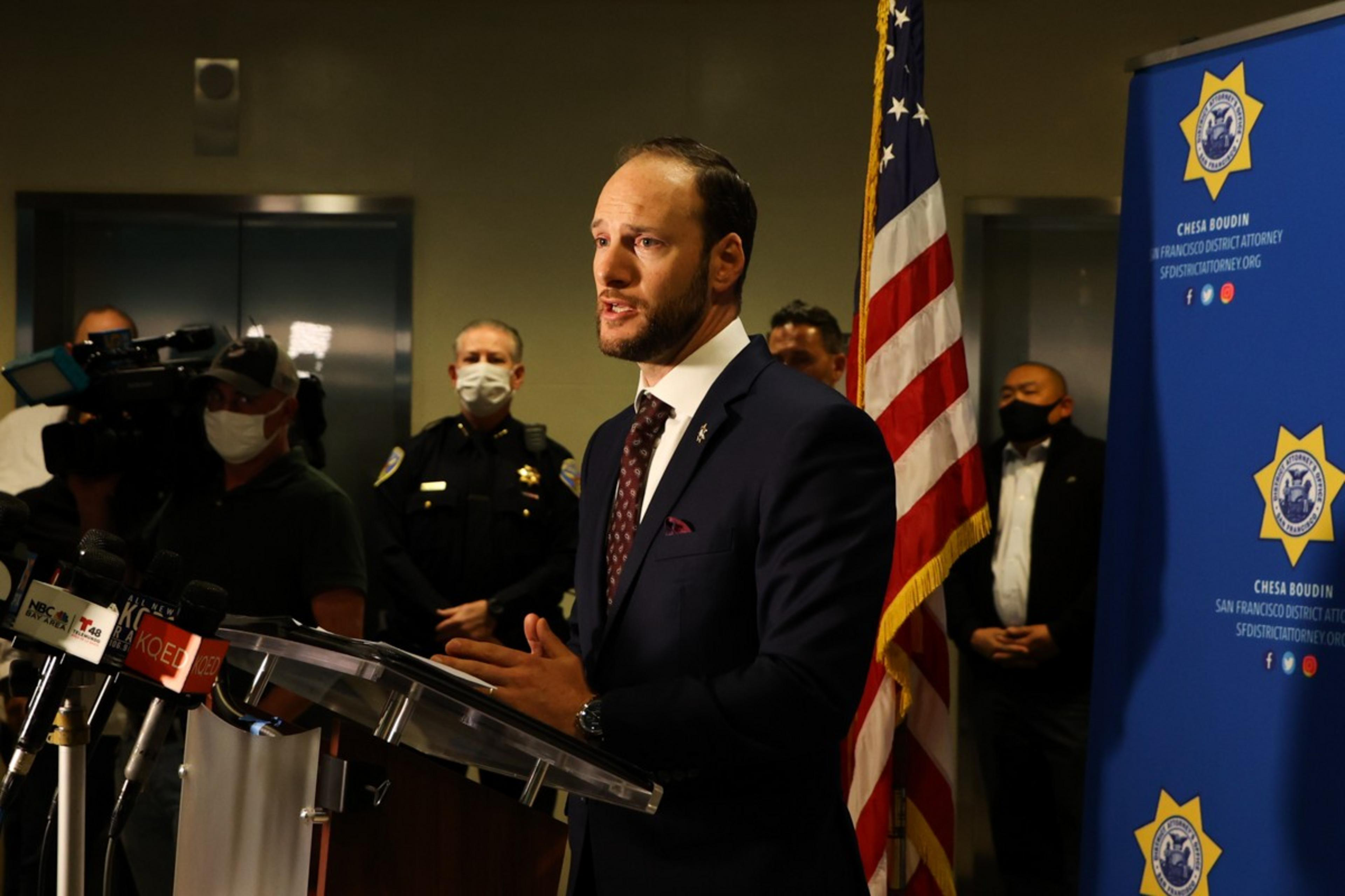 A man in a suit speaks at a podium with multiple microphones. An American flag is beside him. People, including a masked officer, stand behind him.