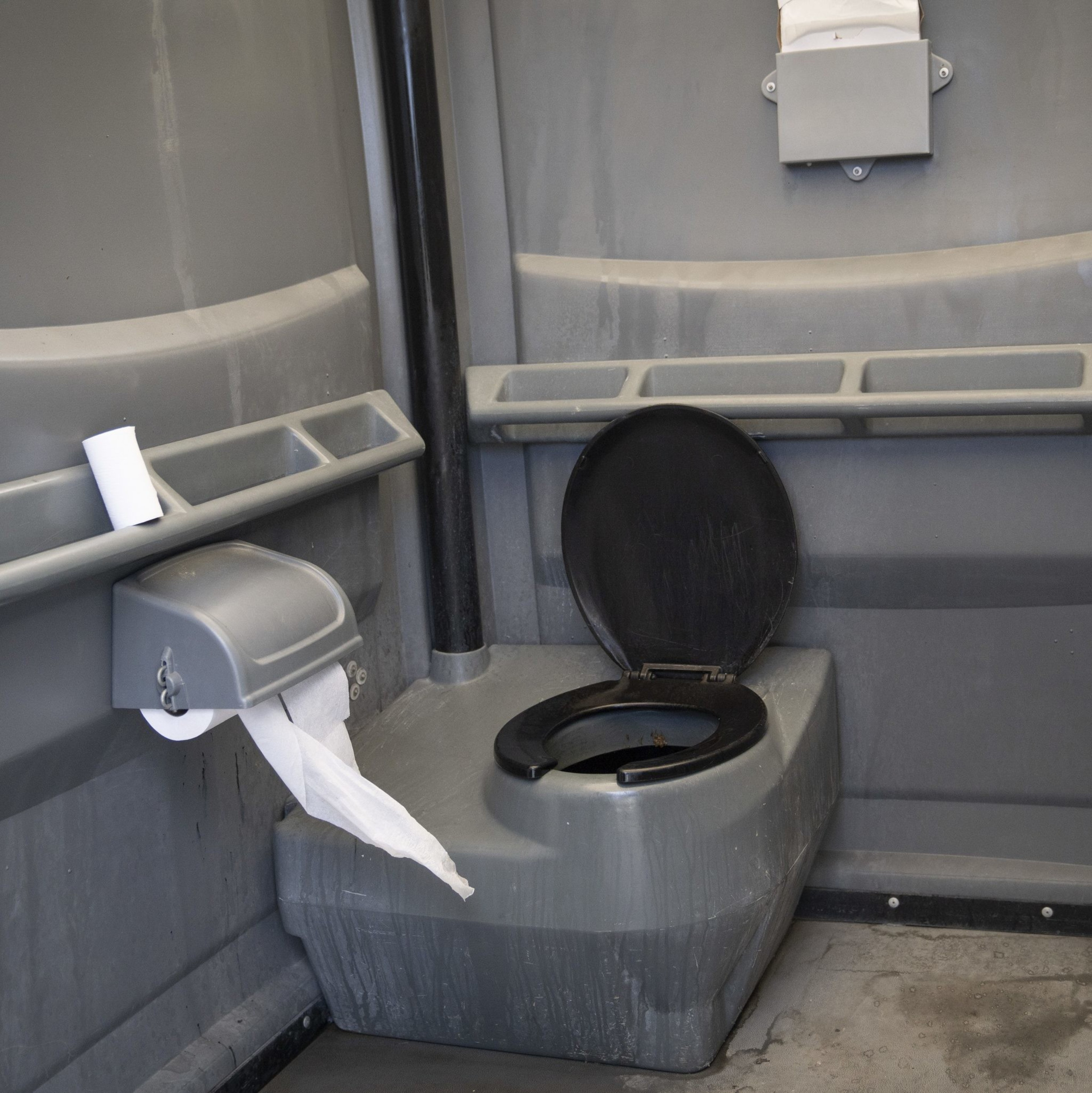 The image shows the interior of a portable toilet with a dark seat, a roll of toilet paper nearby, and some paper protruding from a dispenser.