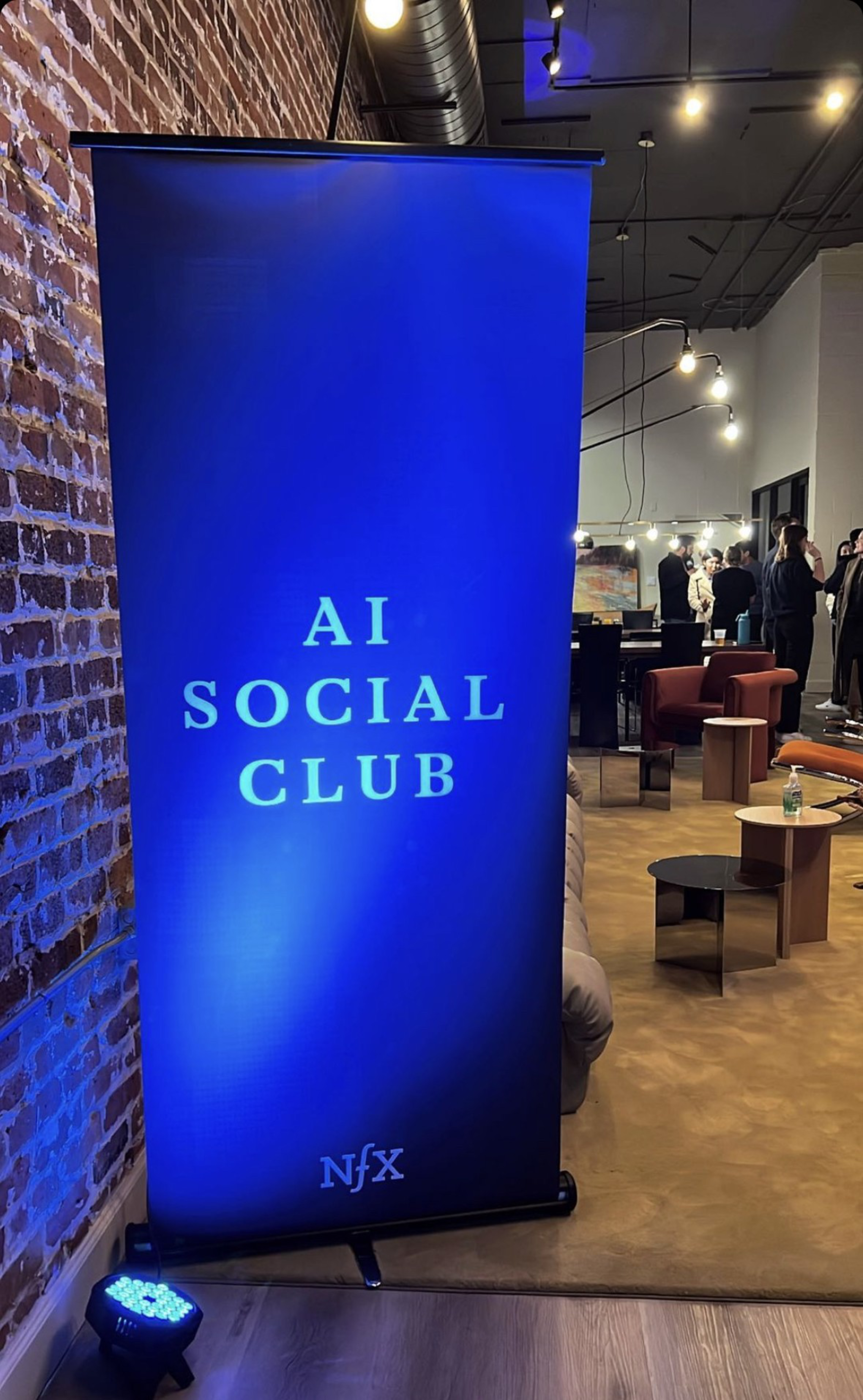 A blue banner with "AI Social Club" and "NFX" stands against a brick wall in a modern lounge where people are mingling.