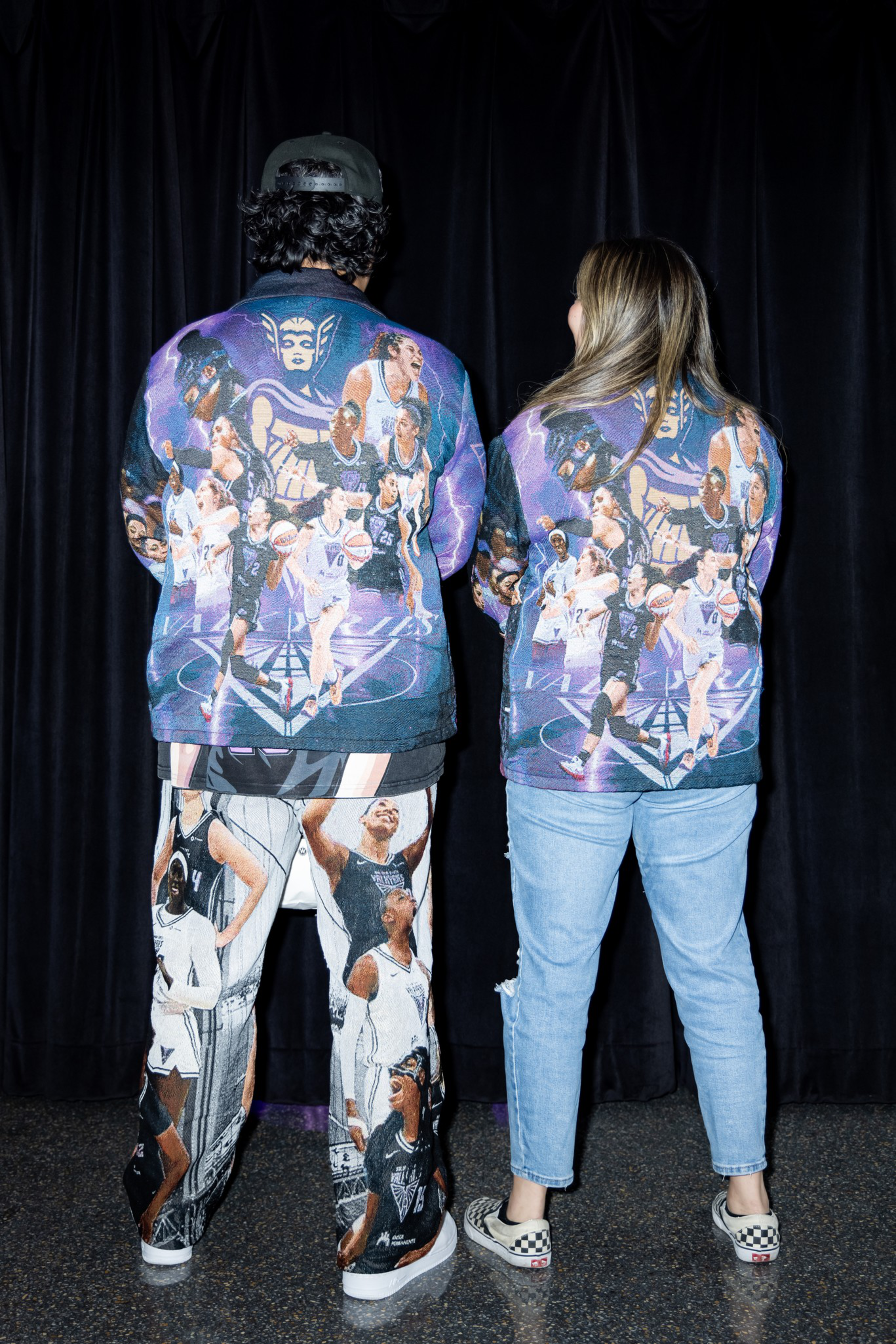 Two people stand with backs facing the camera, wearing matching jackets featuring a vibrant collage of basketball players on a purple background.