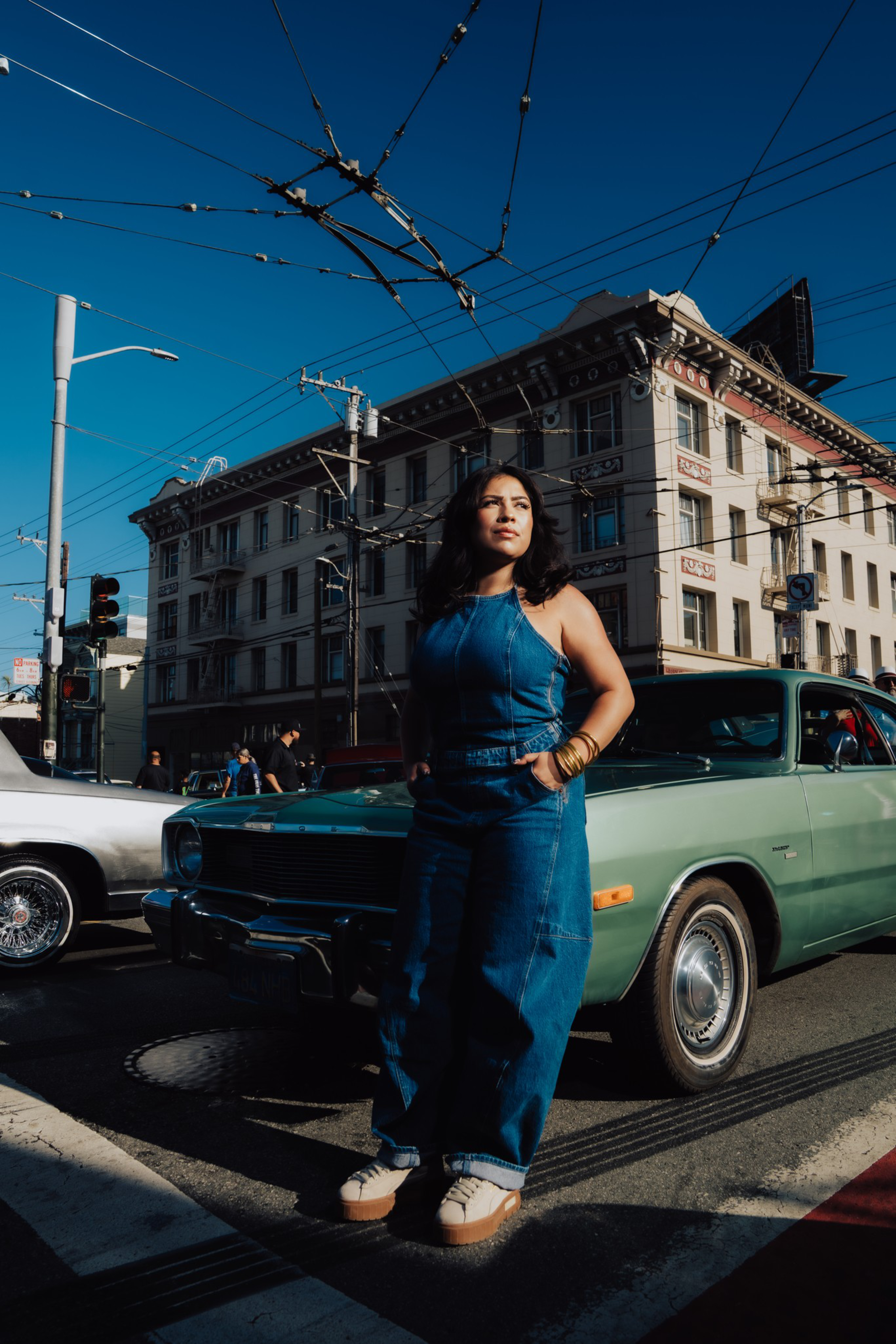 A woman in a blue denim jumpsuit stands confidently on a street corner in front of a green classic car, with a building and power lines behind her.