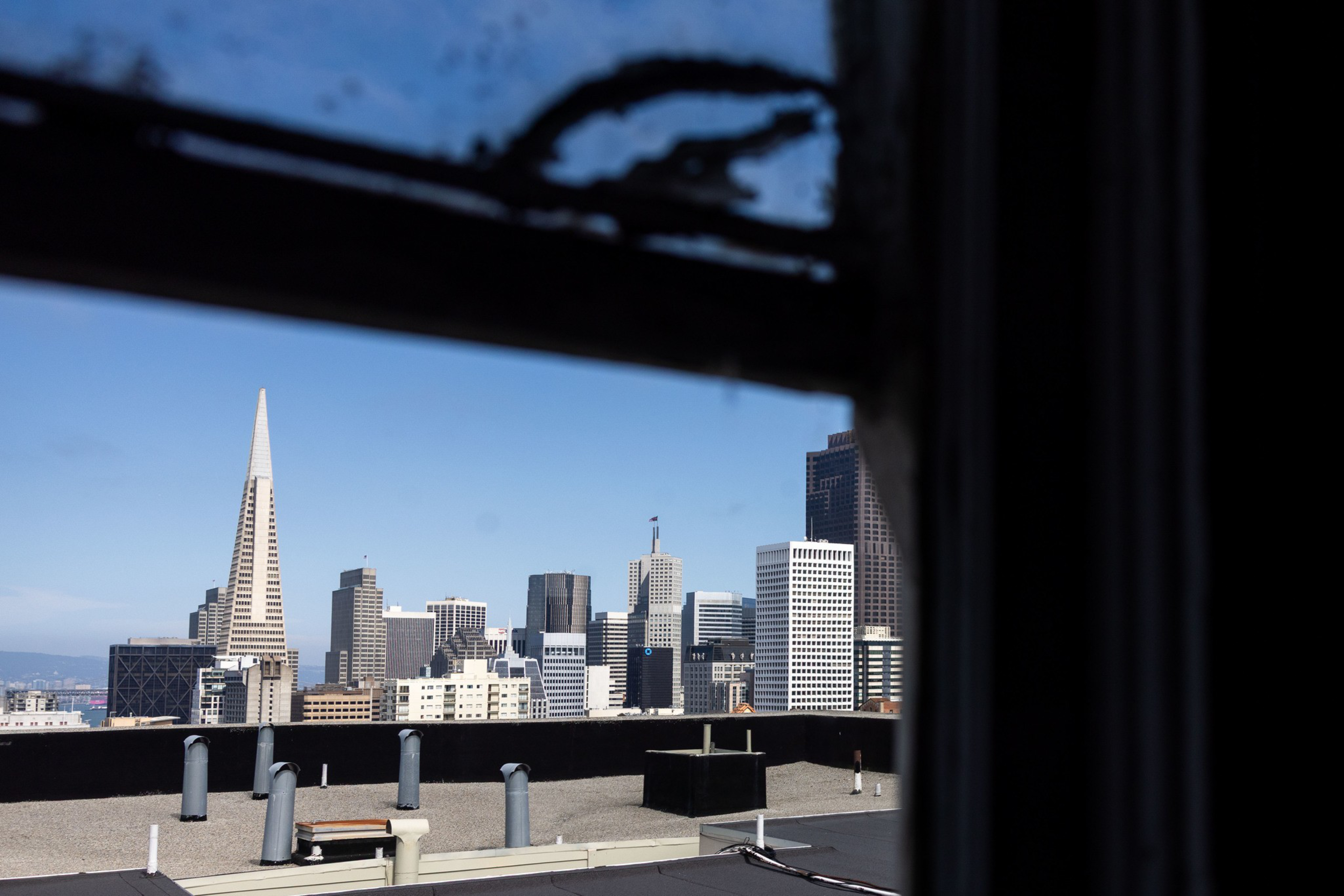 A city skyline with modern skyscrapers, including a tall, pointed white building, is visible through a window with a dark frame and some dirt or wear.