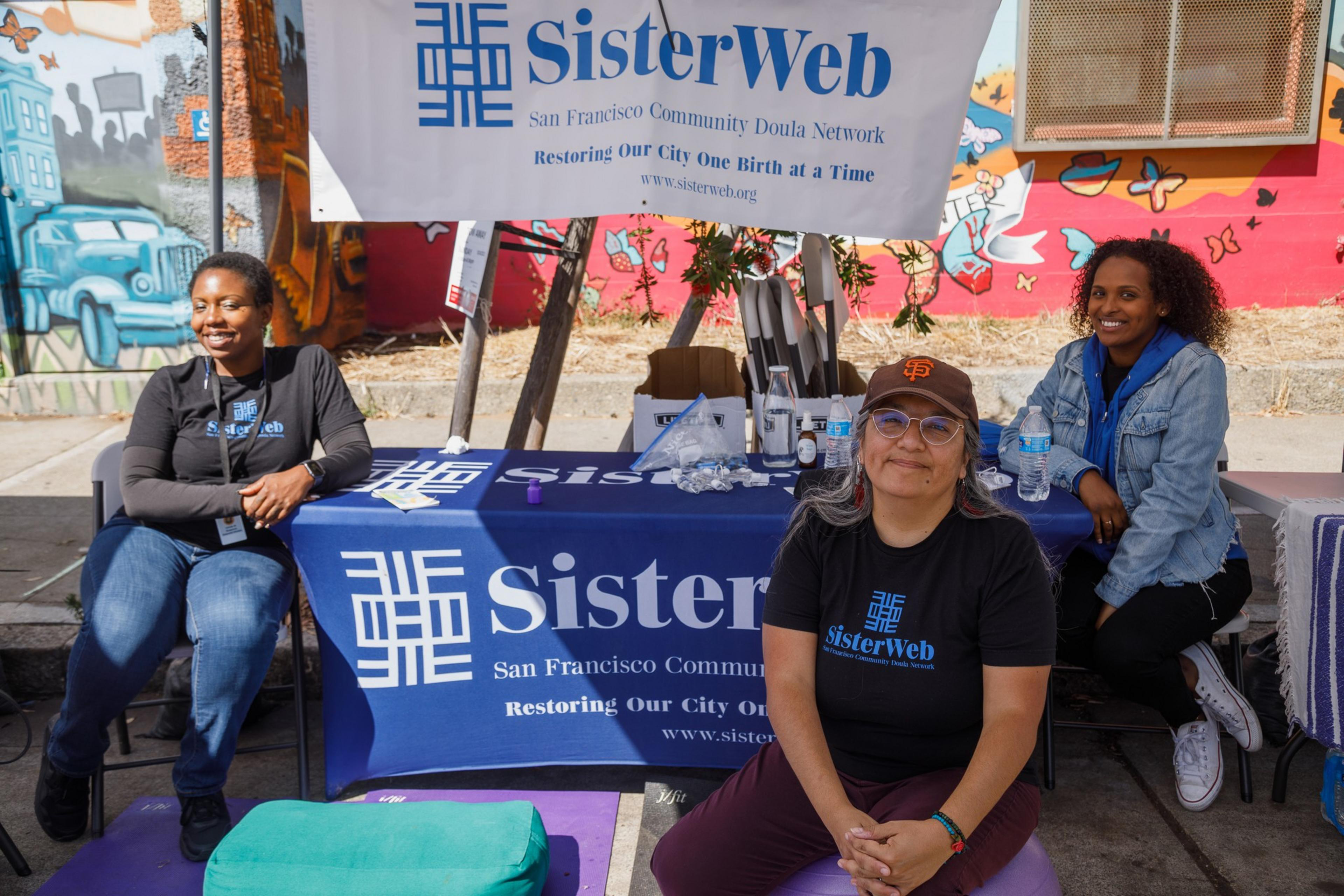 Three individuals sit by a booth with a &quot;SisterWeb&quot; banner, smiling in front of a colorful mural.