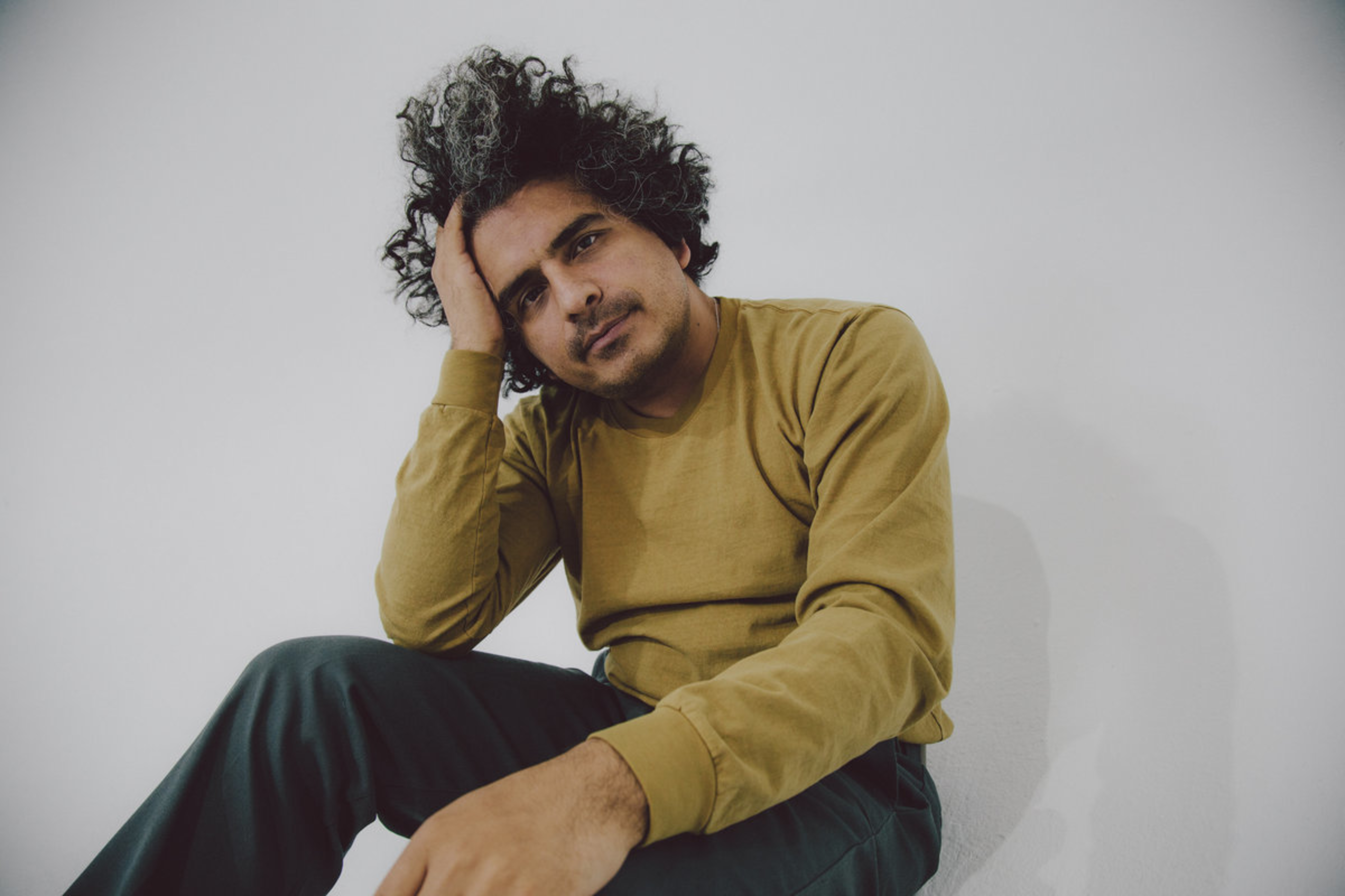 A man with curly hair and a mustache wears a mustard sweater and dark pants, resting his head on one hand while sitting against a white wall.