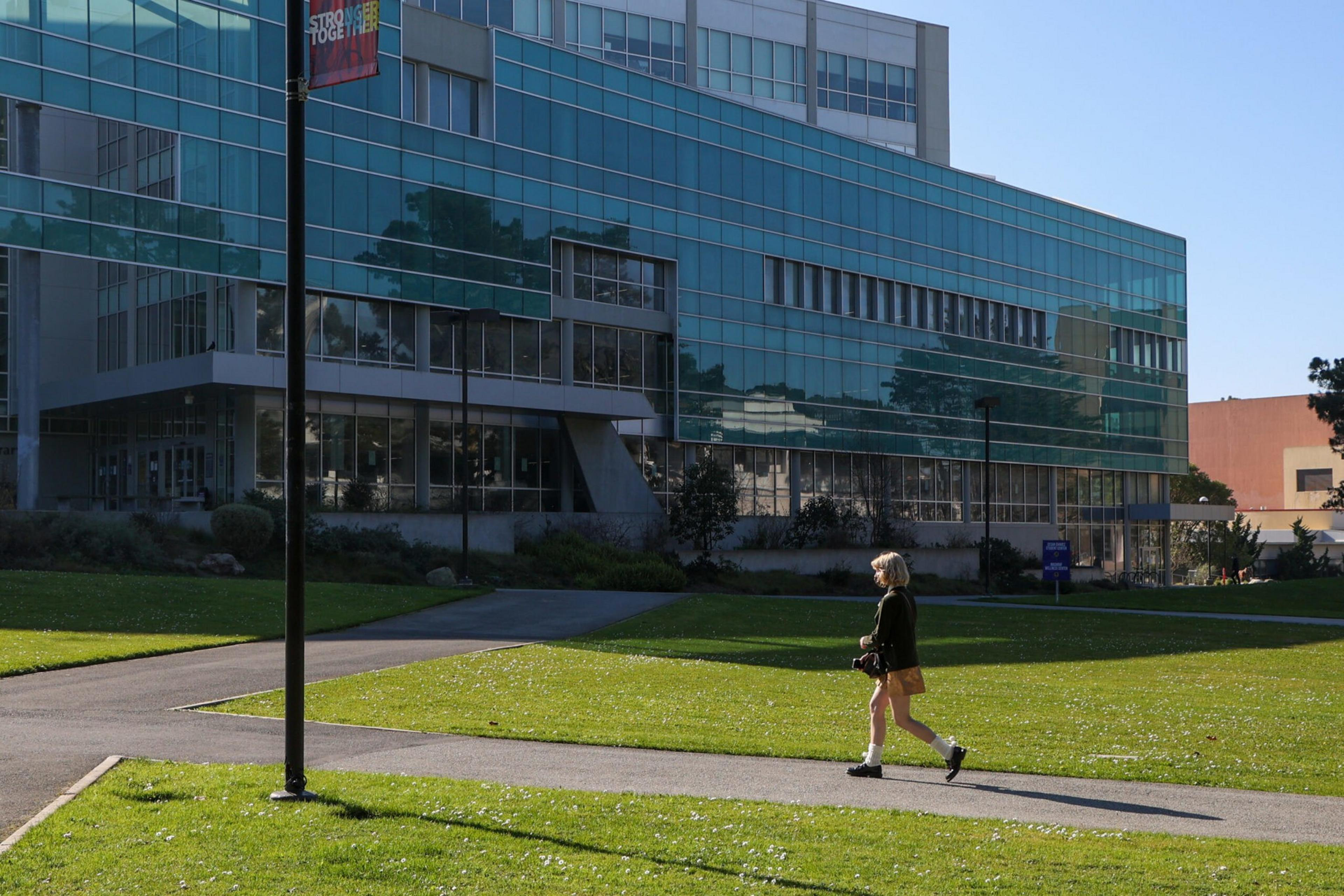 A person walks down a sidewalk toward a large glass-fronted building.