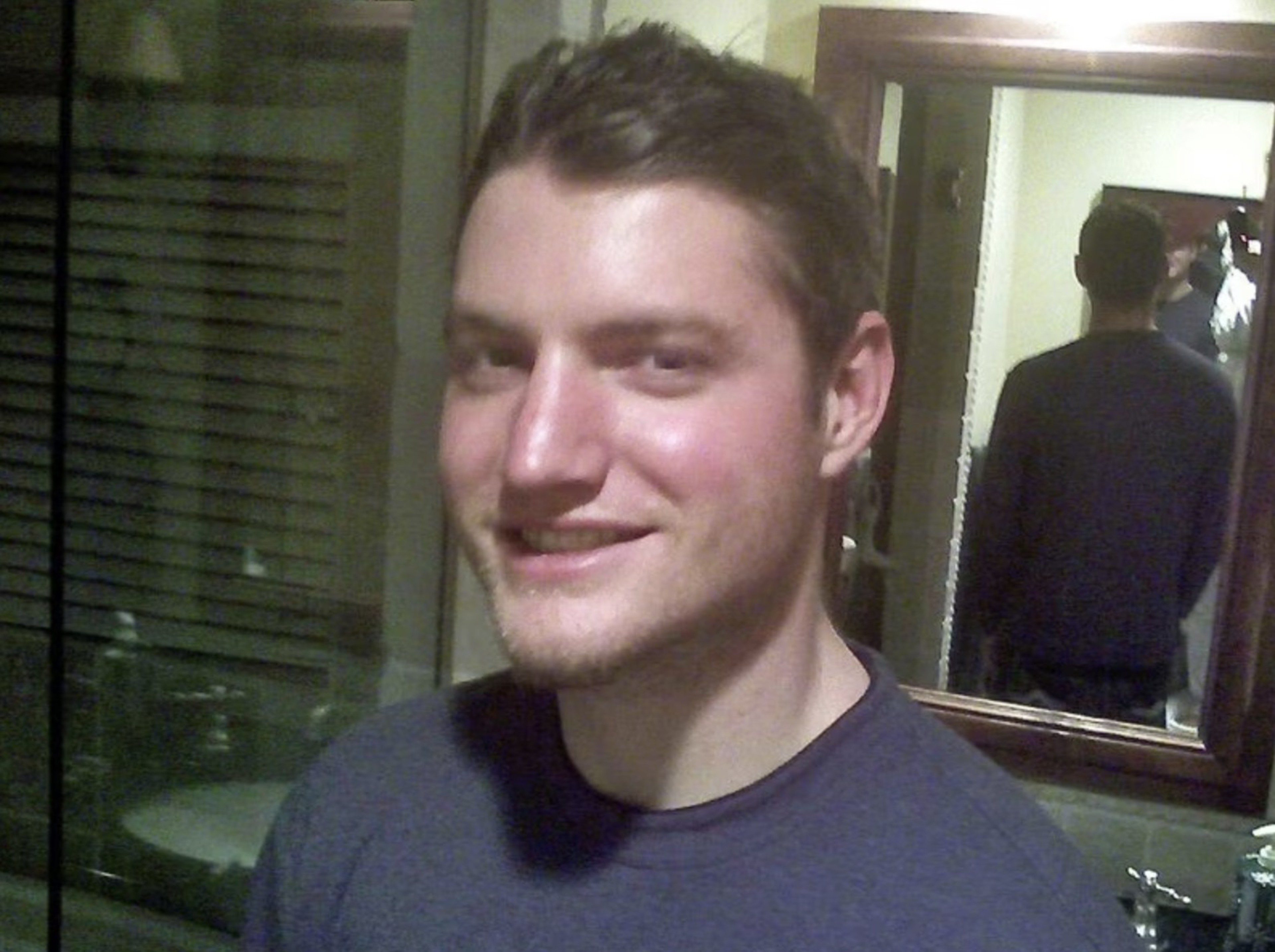 A smiling man with short hair in a blue shirt, indoors, with a reflection in a mirror.