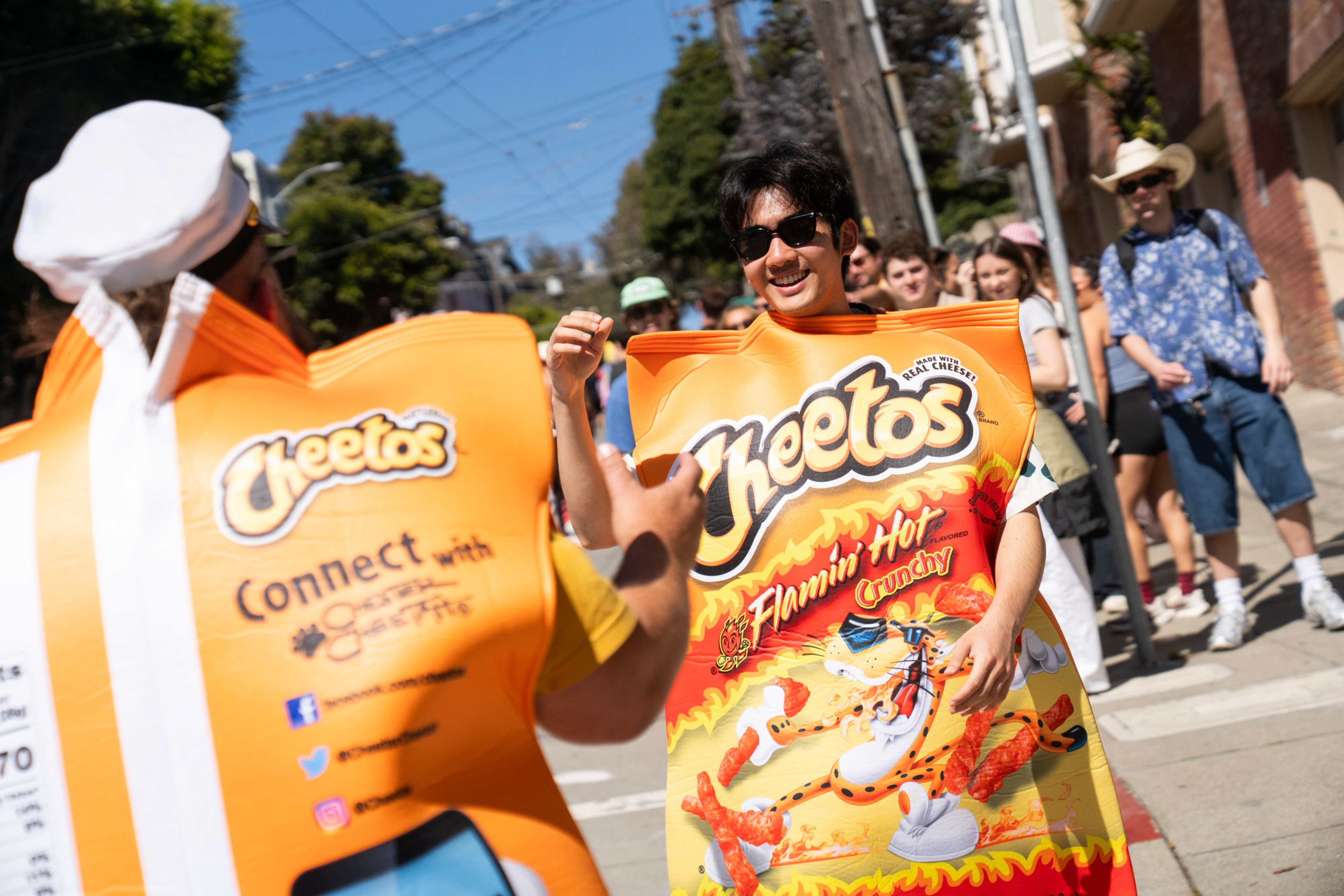 Two people wearing oversized Cheetos Flamin’ Hot Crunchy bags are walking and interacting on a sunny street, with a crowd watching behind them.