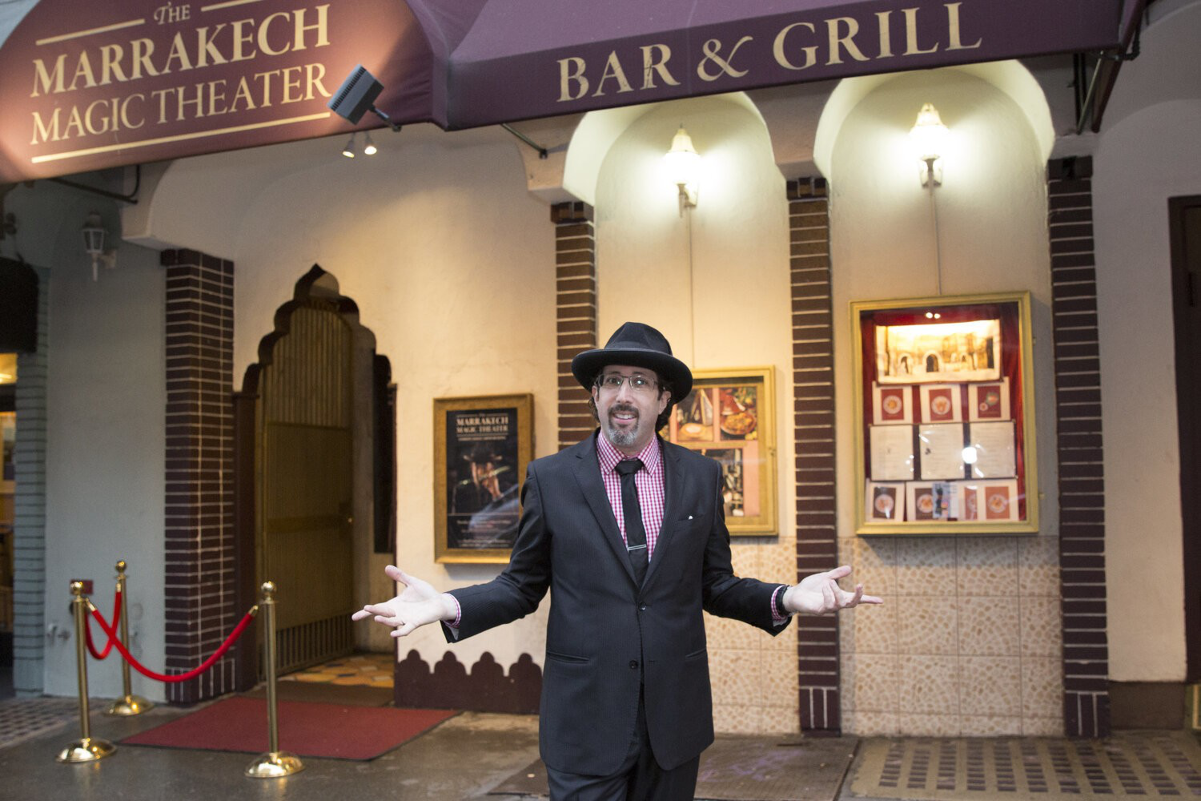 A man in a black suit, hat, and tie stands with arms open in front of The Marrakech Magic Theater bar and grill entrance at night.