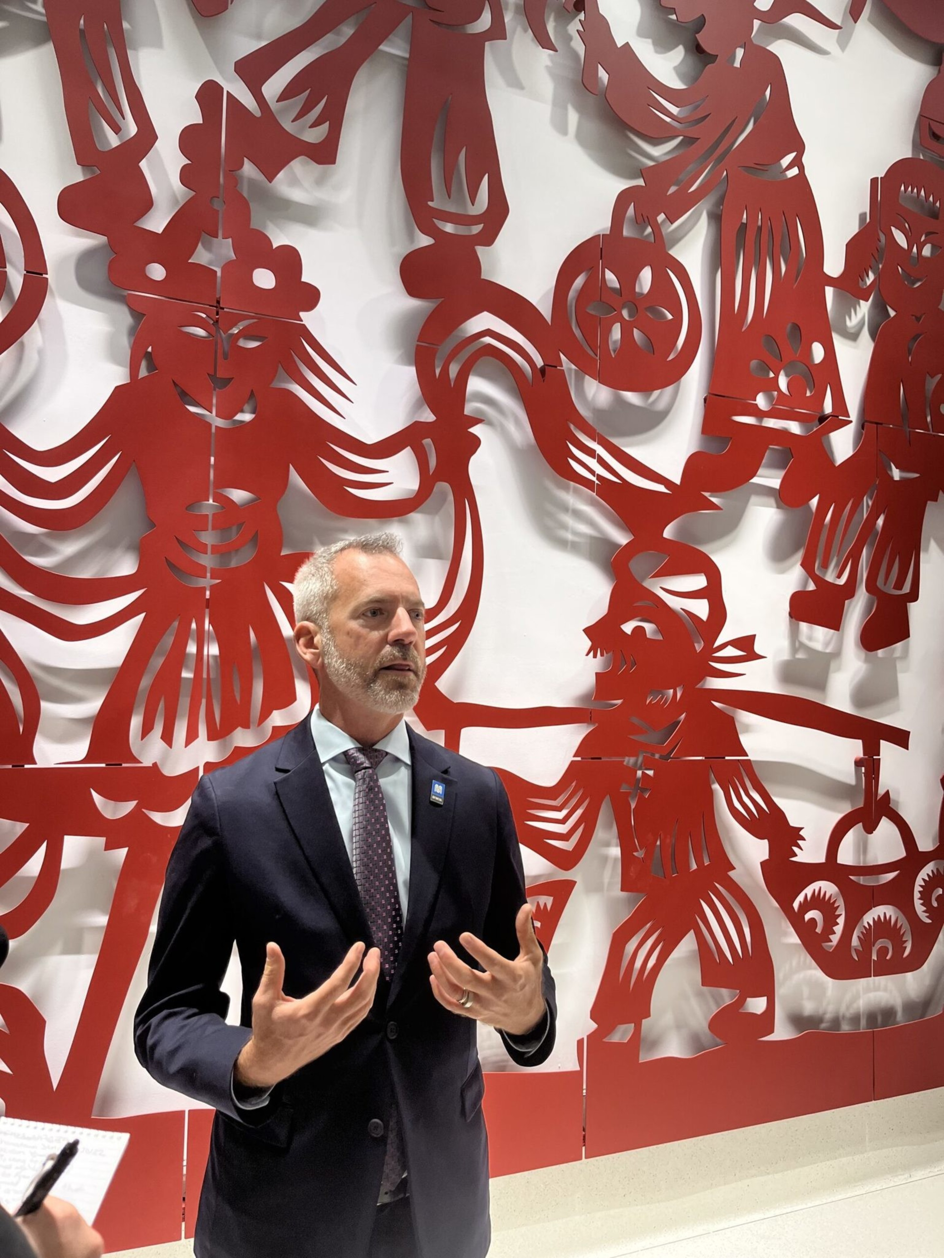 A man in a dark suit and tie gestures while speaking in front of a large, intricate red wall mural with stylized human figures.