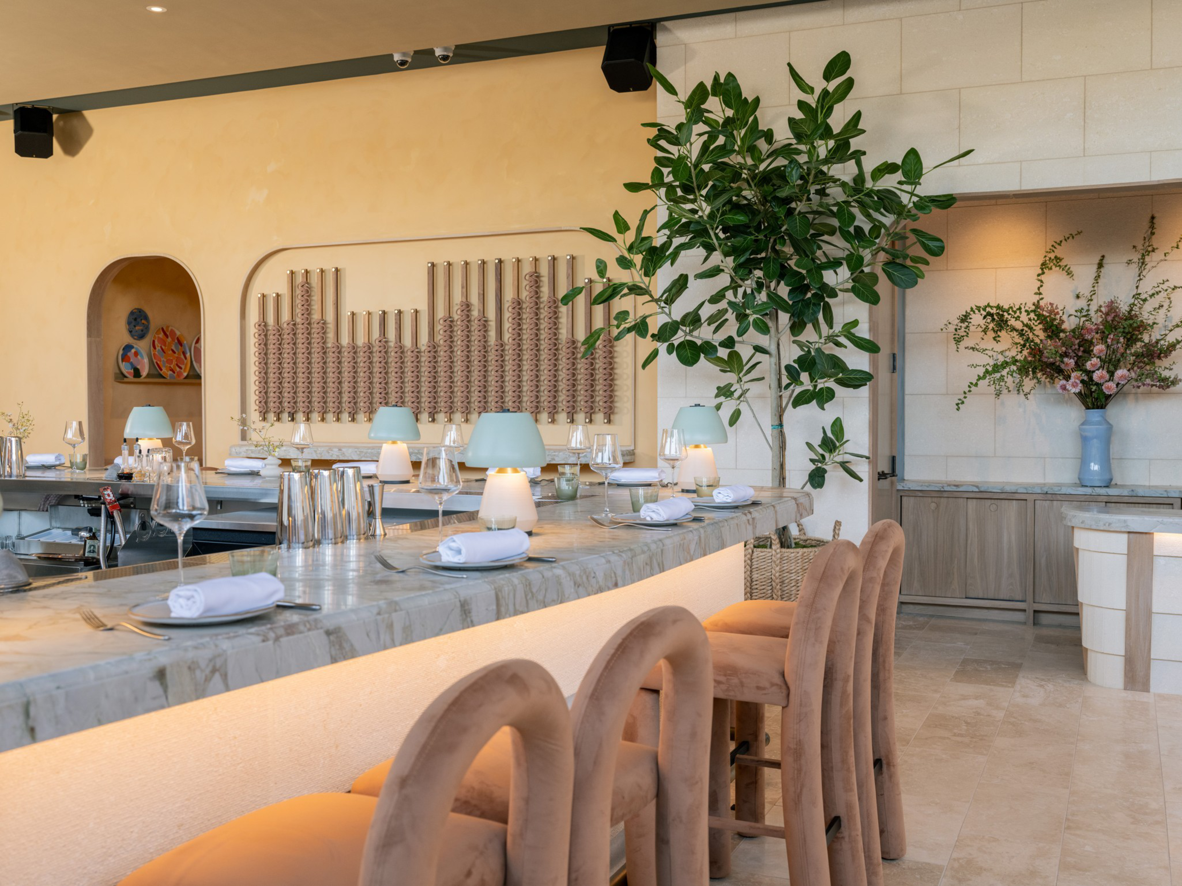 A marble countertop bar with beige upholstered stools is set with white napkins, glassware, blue lamps, and surrounded by plants and modern decor.