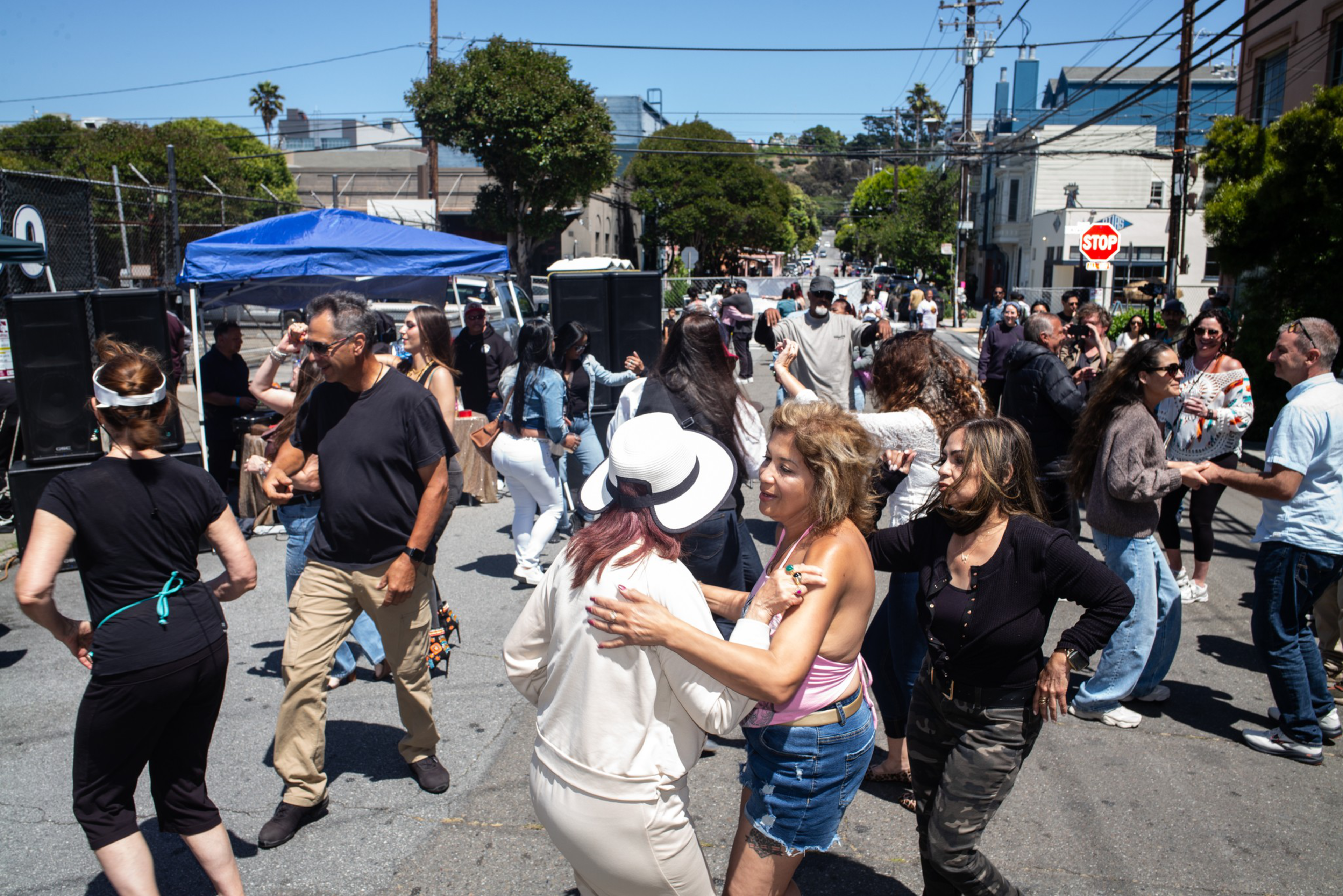 People of various ages dance and socialize in a sunlit street festival with a blue canopy and speakers in the background.