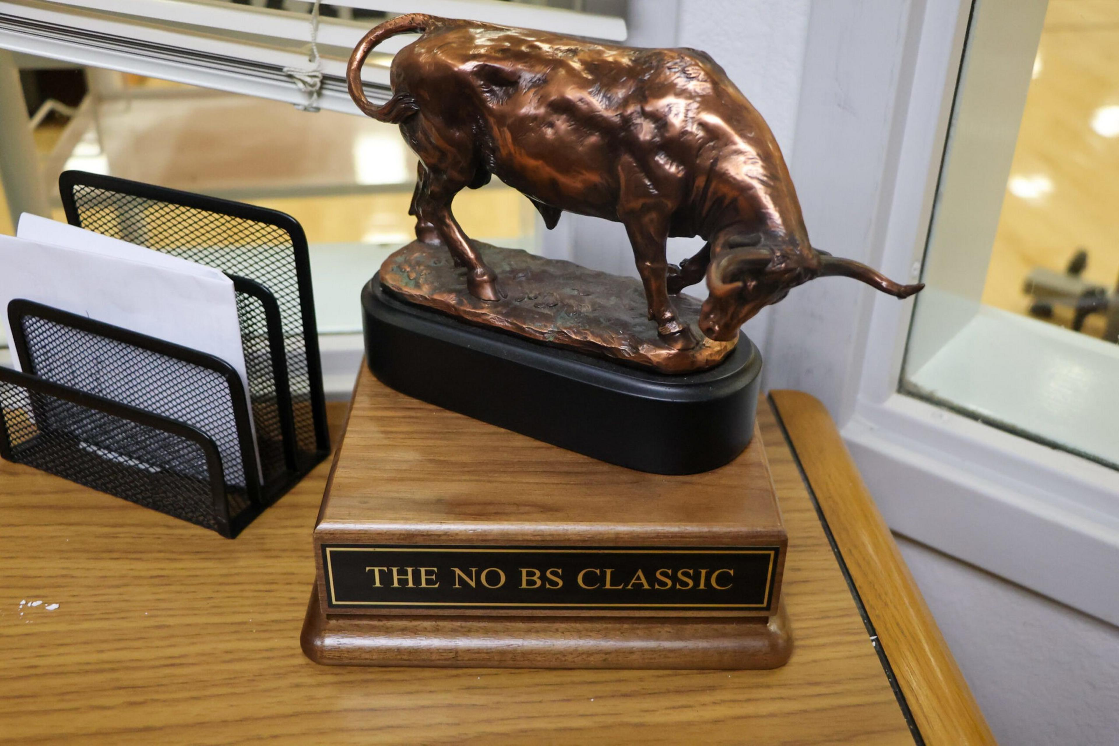 A bronze bull statue stands on a wooden base with a plaque reading "THE NO BS CLASSIC," placed on a wooden desk next to a black mesh organizer.