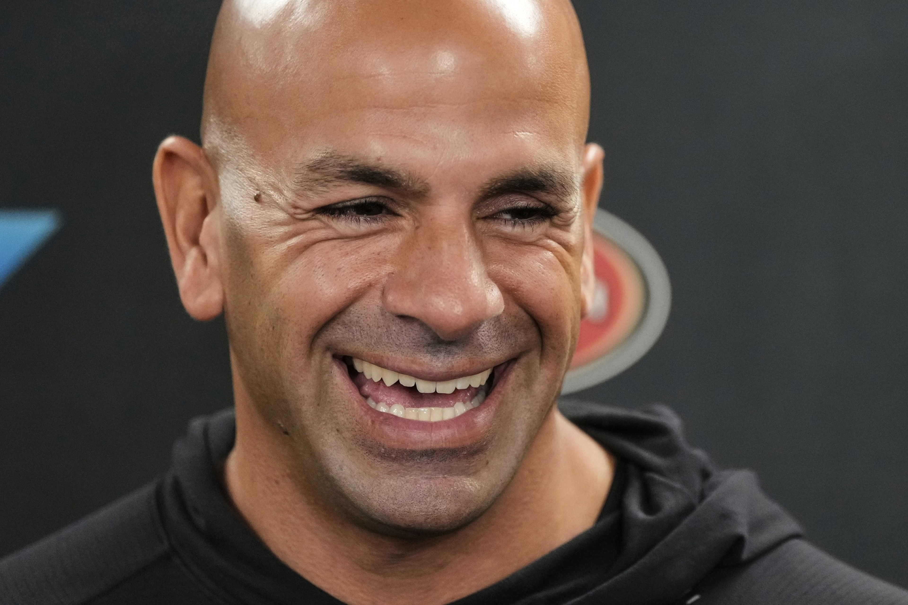 A man with a bald head and a broad smile is wearing a black hoodie. He has deep laugh lines, and there's a blurred logo in the background.