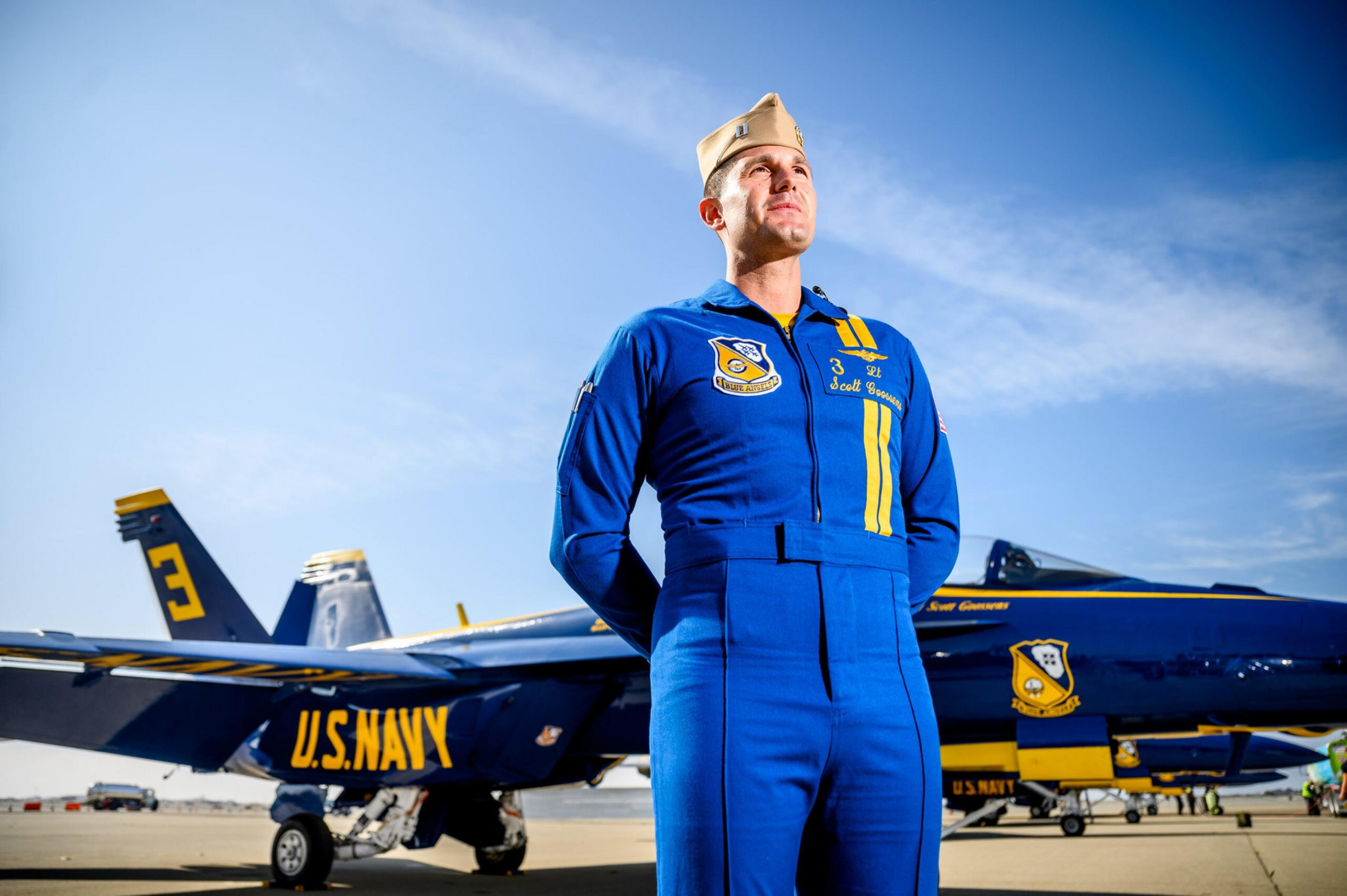 A U.S. Navy pilot in a blue flight suit and tan cap stands confidently with arms behind his back in front of a Blue Angels jet numbered 3 on a sunny day.