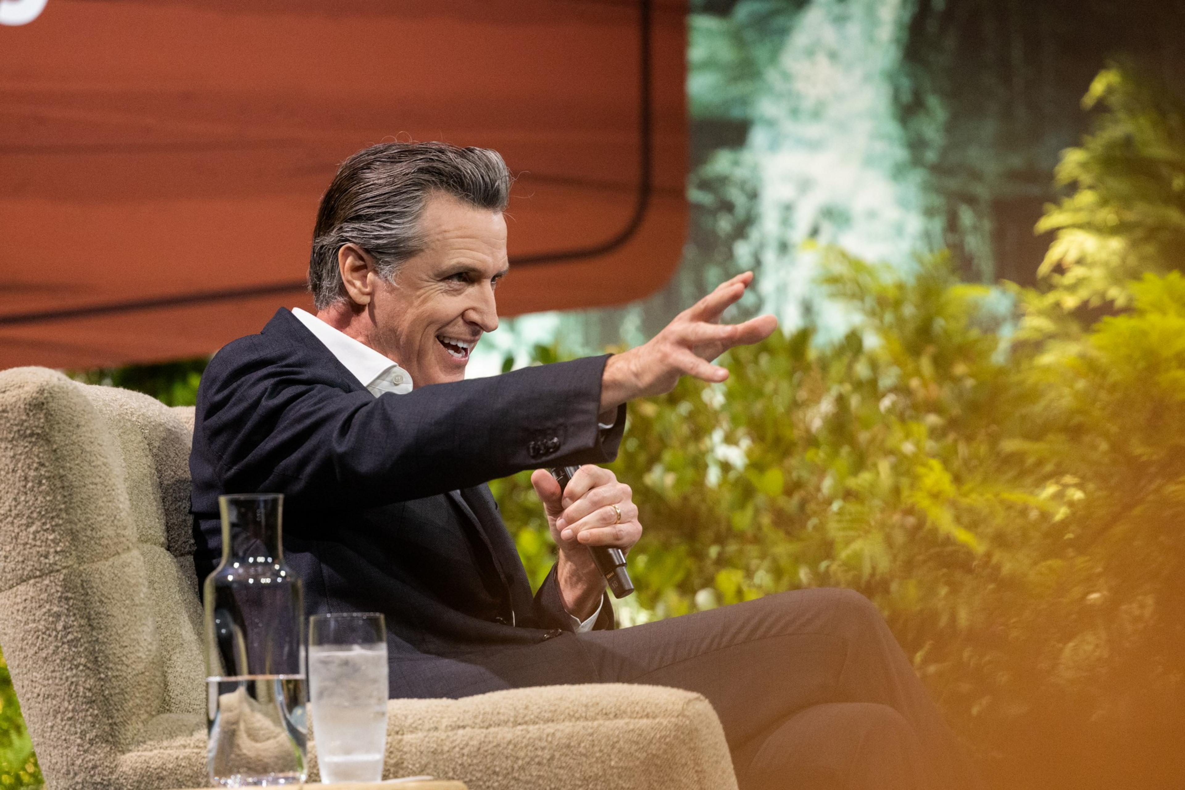 A man in a dark suit sits on a beige chair, holding a microphone in one hand and gesturing with the other, smiling during a conversation.
