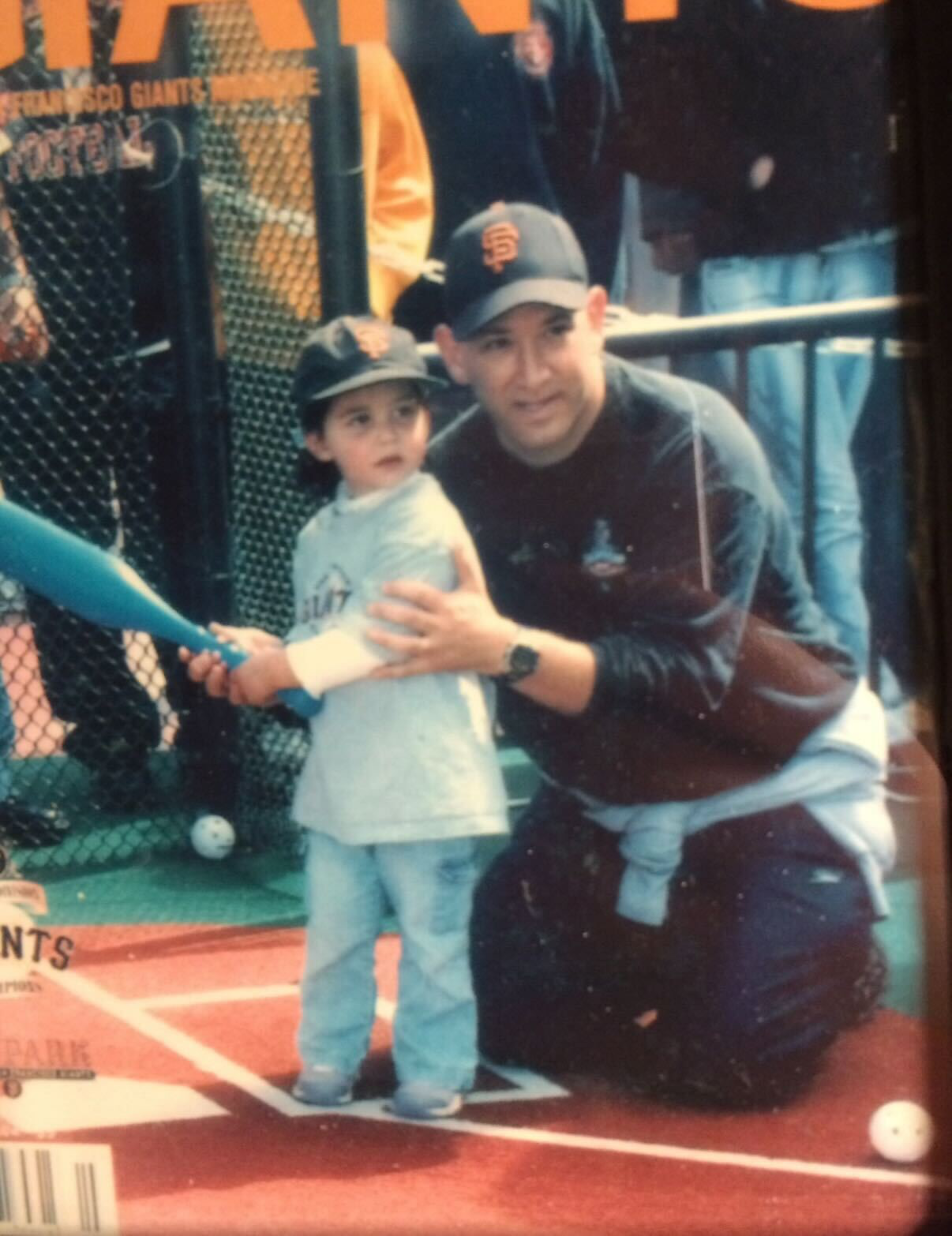 A man kneels behind a young child, guiding the child holding a blue baseball bat at a batting cage, both wearing San Francisco Giants caps.