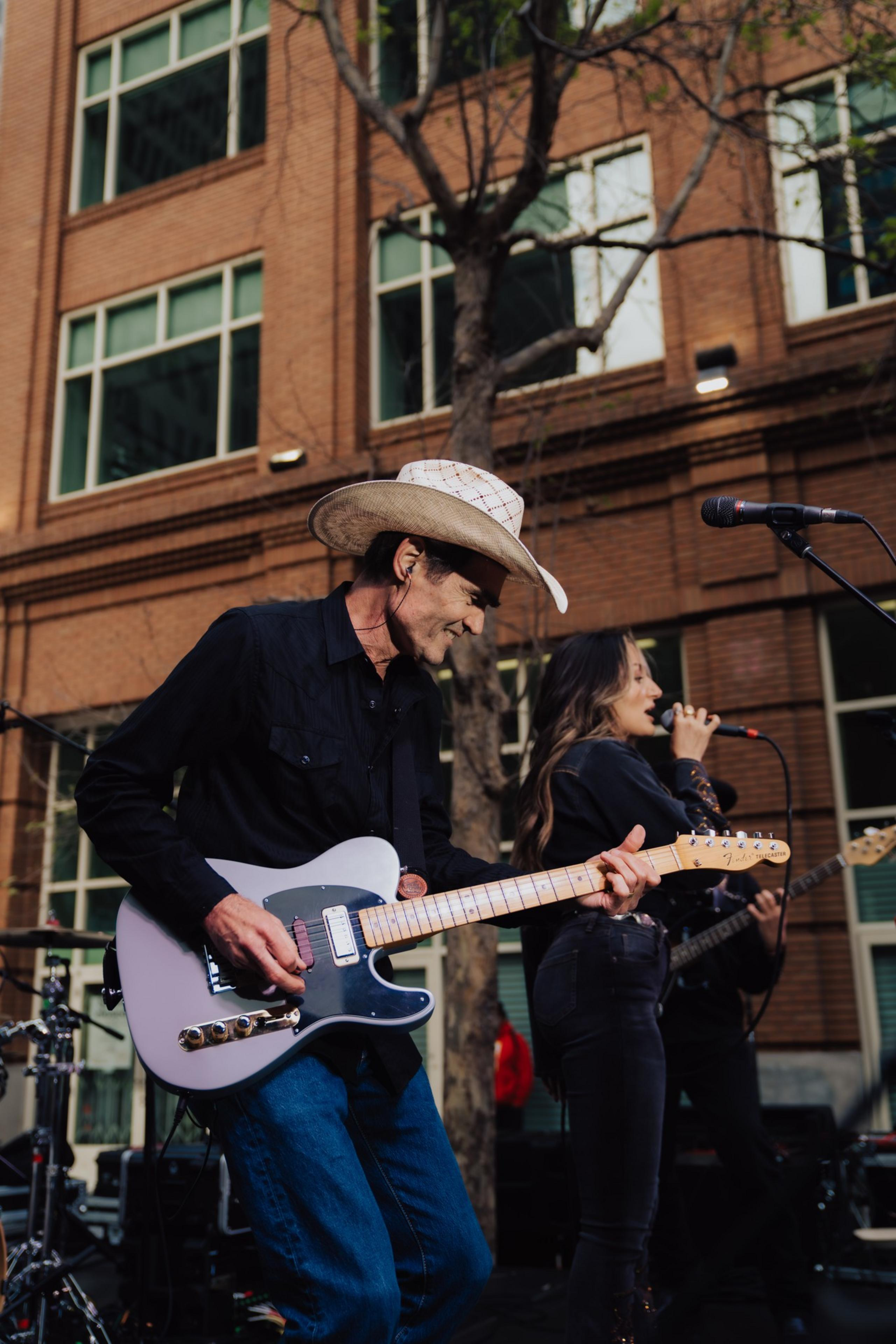 A man in a cowboy hat plays an electric guitar on an outdoor stage, while a woman sings beside him. They are in front of a brick building with large windows.