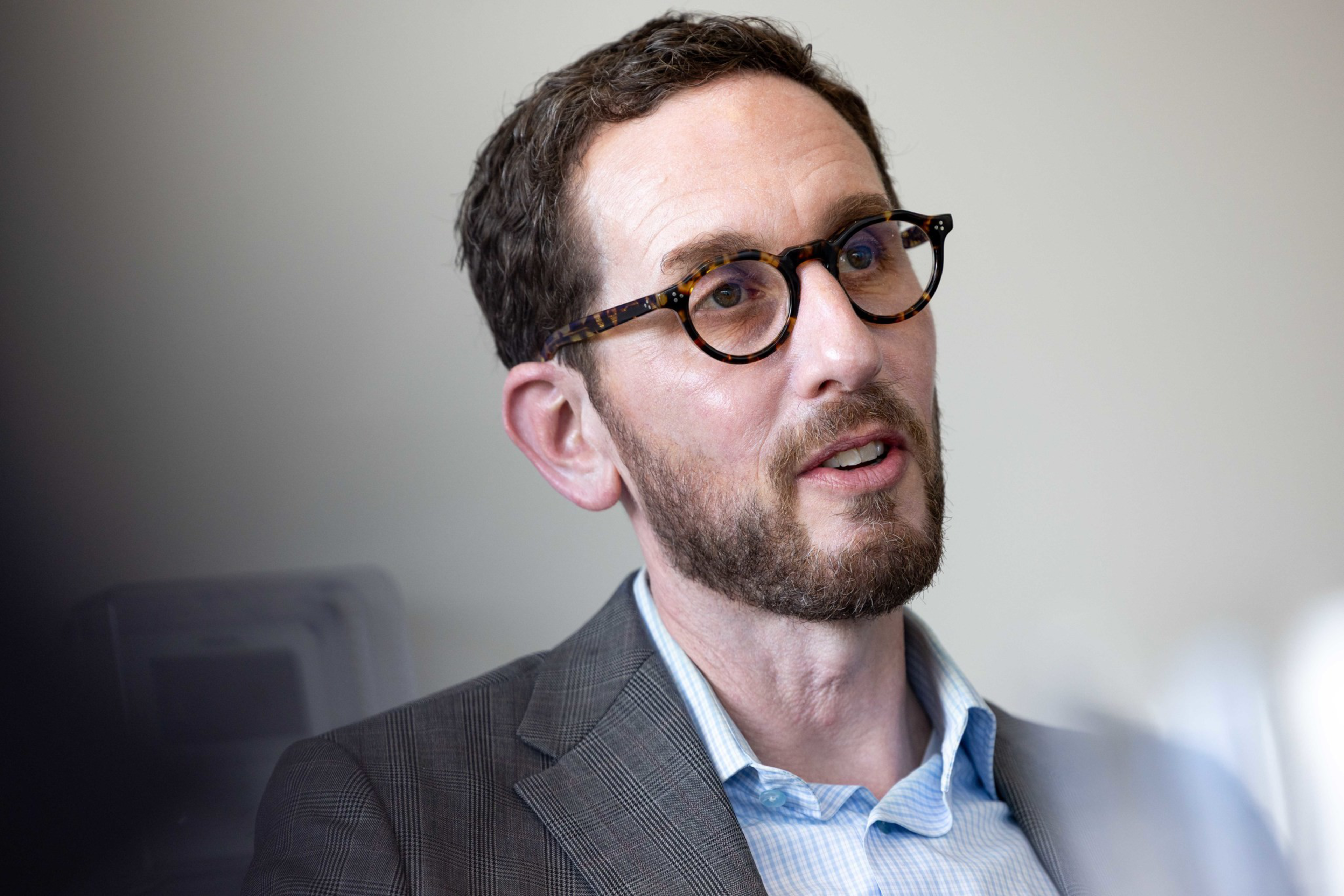 A man with short dark hair, a beard, round tortoiseshell glasses, and a plaid blazer over a light blue shirt is speaking or listening.