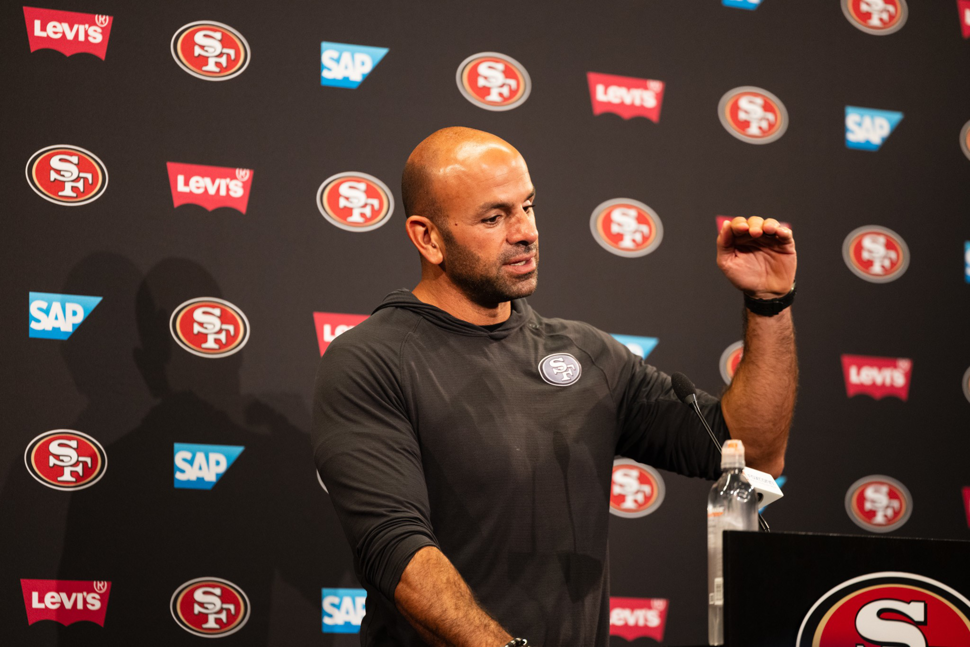 A man in a black San Francisco 49ers hoodie speaks at a podium with logos of 49ers, Levi's, and SAP in the background.