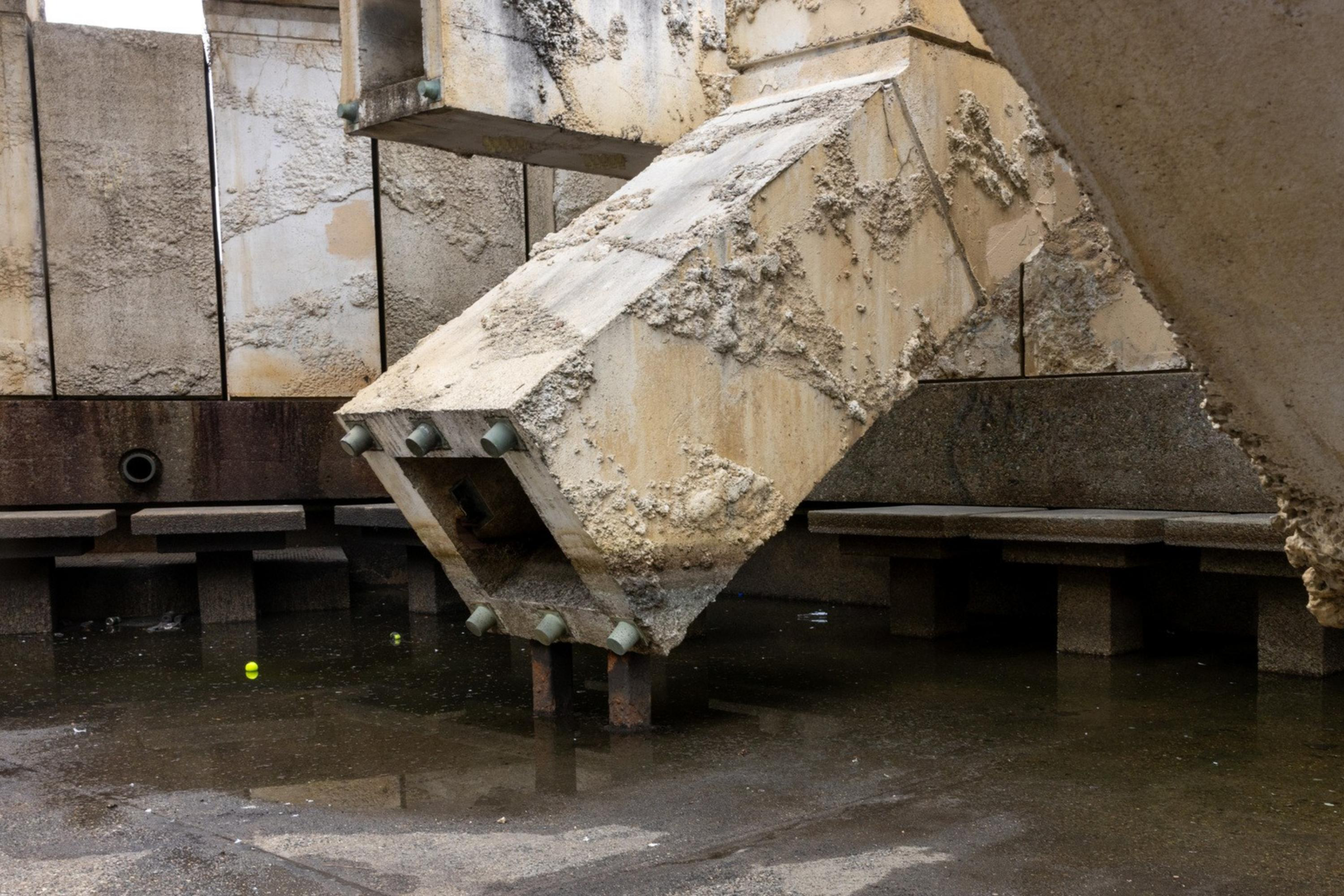 A rugged, weathered concrete structure over a reflective, wet ground with benches in the background. A small yellow ball is visible on the ground.